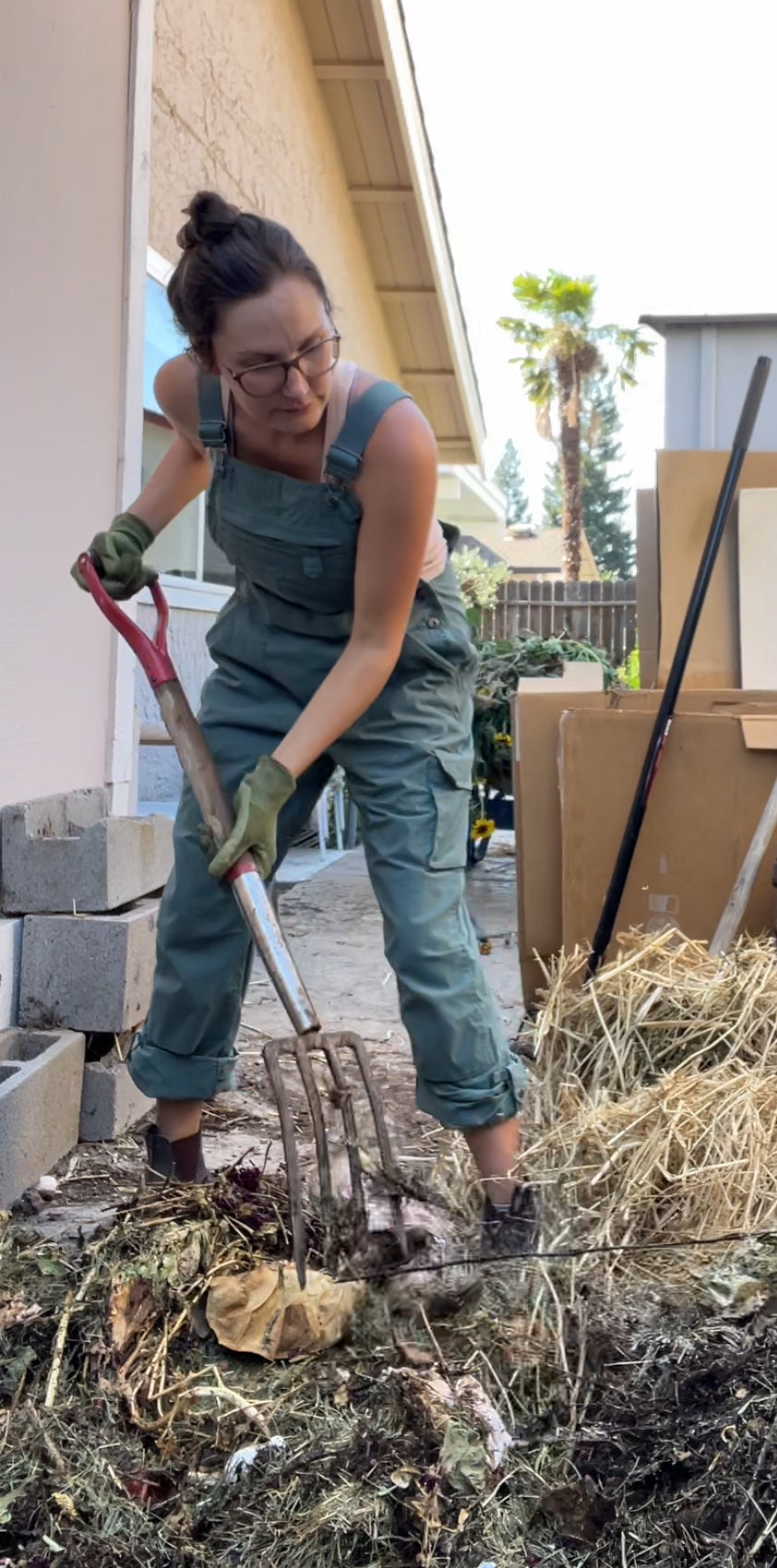 A woman wearing glasses and green overalls uses a pitchfork to turn a compost pile outdoors, surrounded by straw, cardboard, and cinder blocks. She is working in a backyard near a beige house.