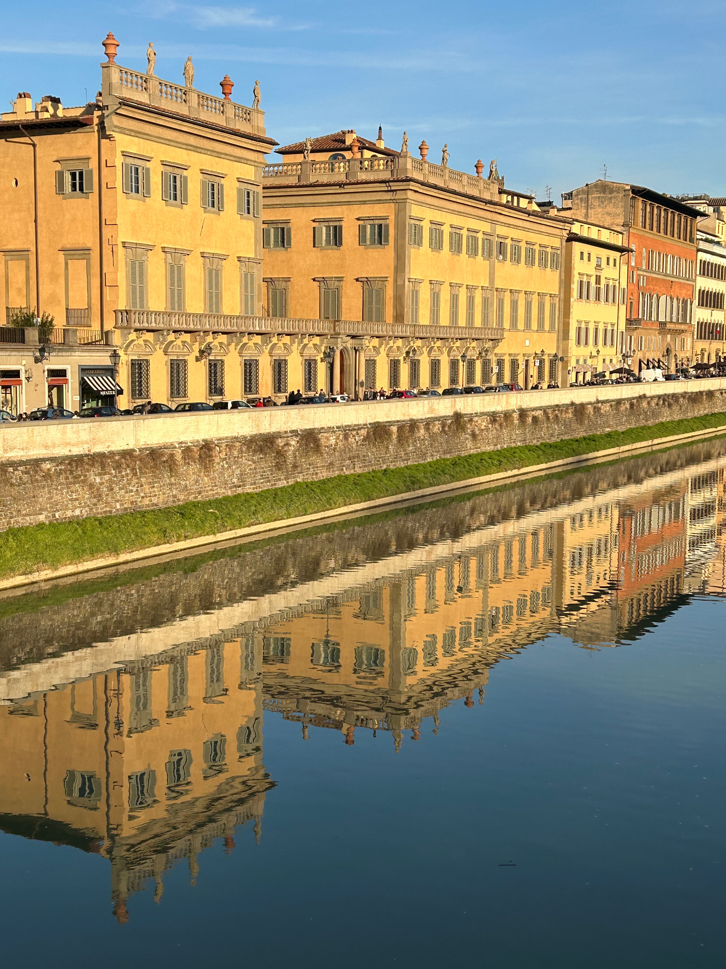 Row of historic yellow buildings with ornate rooftops reflected clearly in the calm river below, under a blue sky. A stone embankment separates the buildings from the water.