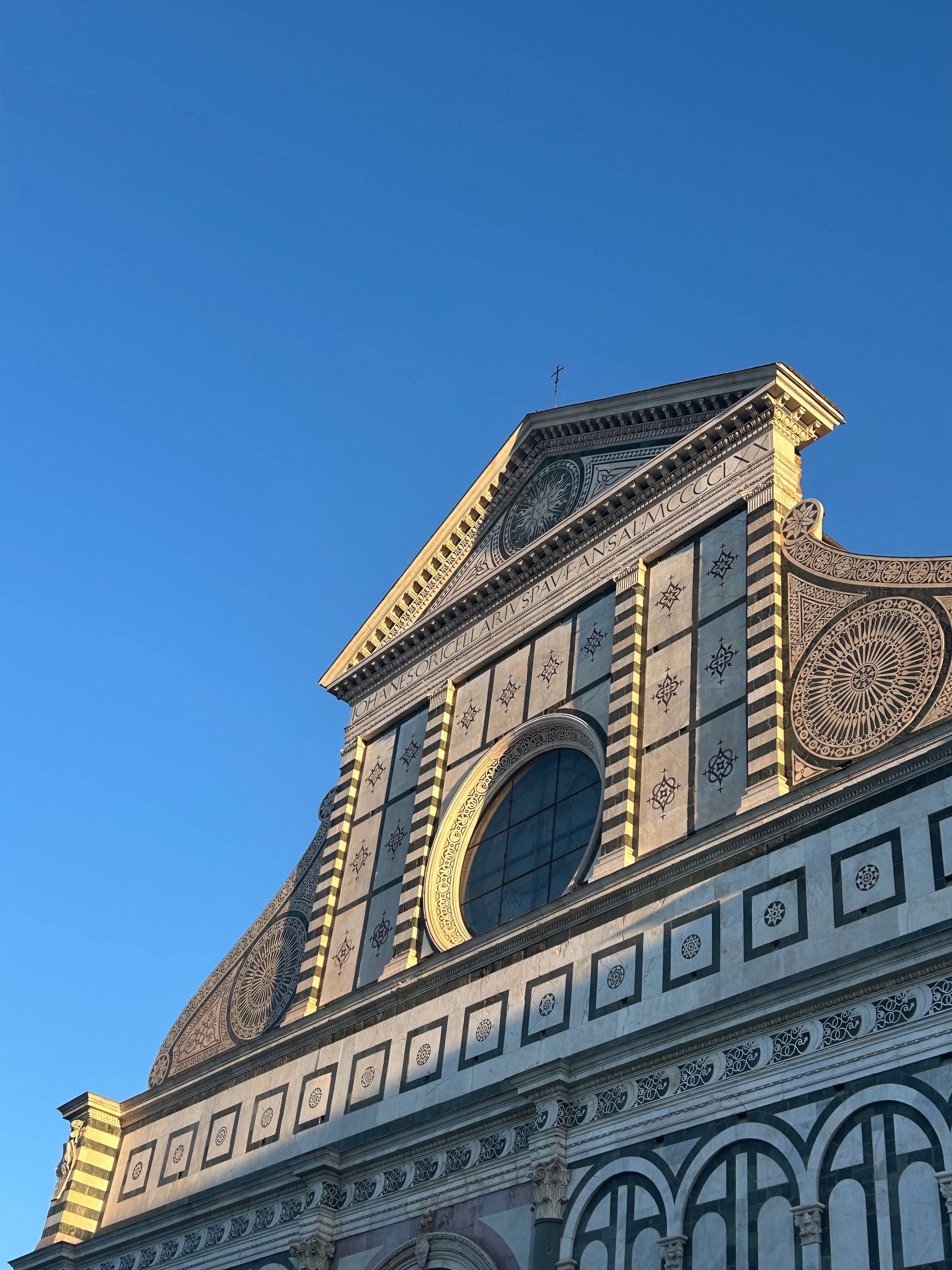 The upper facade of a historic church with ornate geometric designs and a large circular window, lit by sunlight against a clear blue sky.