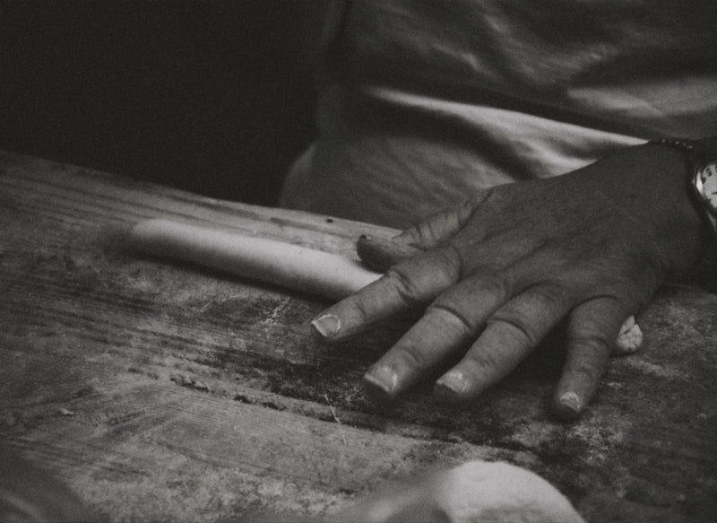 A close-up, black-and-white image of a person’s hand rolling dough on a wooden surface, suggesting bread or pastry preparation. The scene captures texture and focus on the hand’s motion.