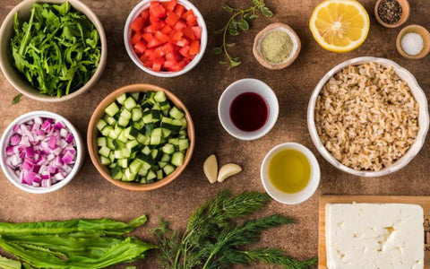 A variety of fresh salad ingredients in bowls, including chopped tomatoes, cucumbers, red onions, cooked brown rice, leafy greens, feta cheese, lemon, olive oil, garlic, herbs, and spices on a brown surface.