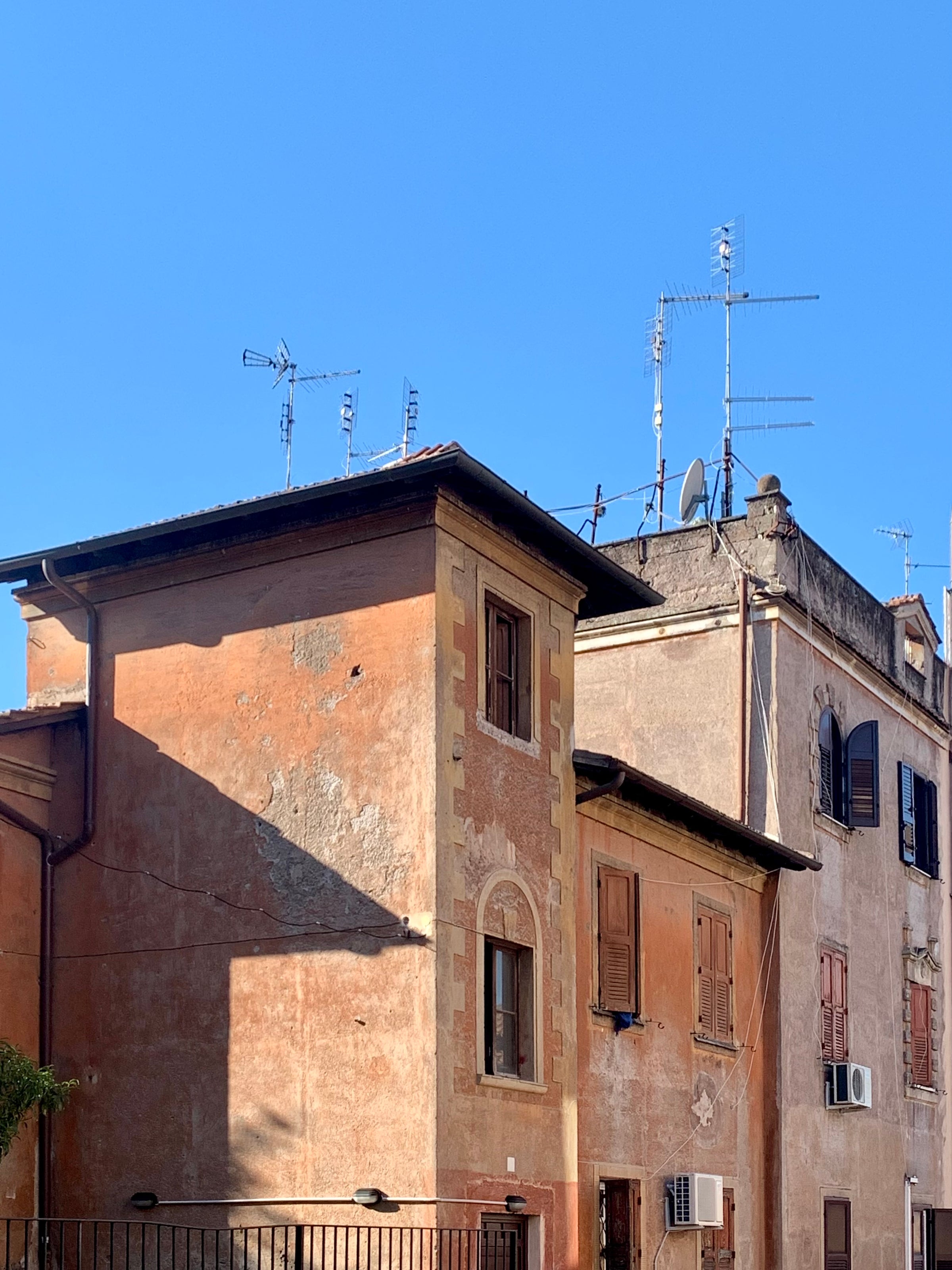 Old, weathered buildings with terracotta walls and wooden shutters stand under a clear blue sky. Several TV antennas and satellite dishes are mounted on the rooftops. Shadows fall across the walls.