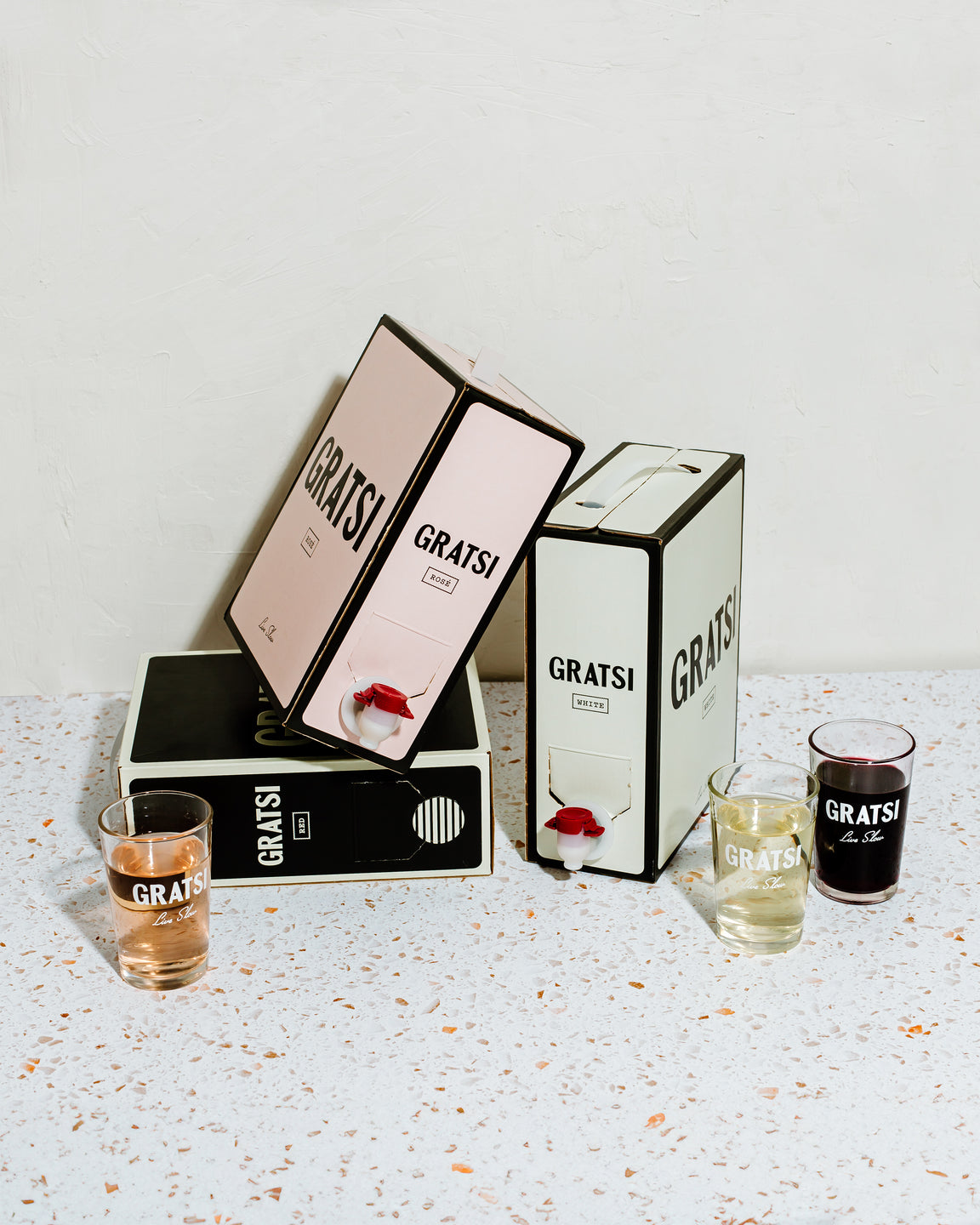 Three box wine containers labeled GRATSI in pink, white, and black are arranged on a terrazzo surface. Each box has a spout, with three glasses in front, each filled with different colored wine.