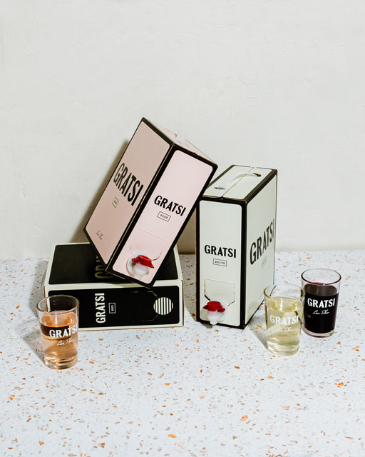 Three box wine containers labeled GRATSI in pink, white, and black are arranged on a terrazzo surface. Each box has a spout, with three glasses in front, each filled with different colored wine.