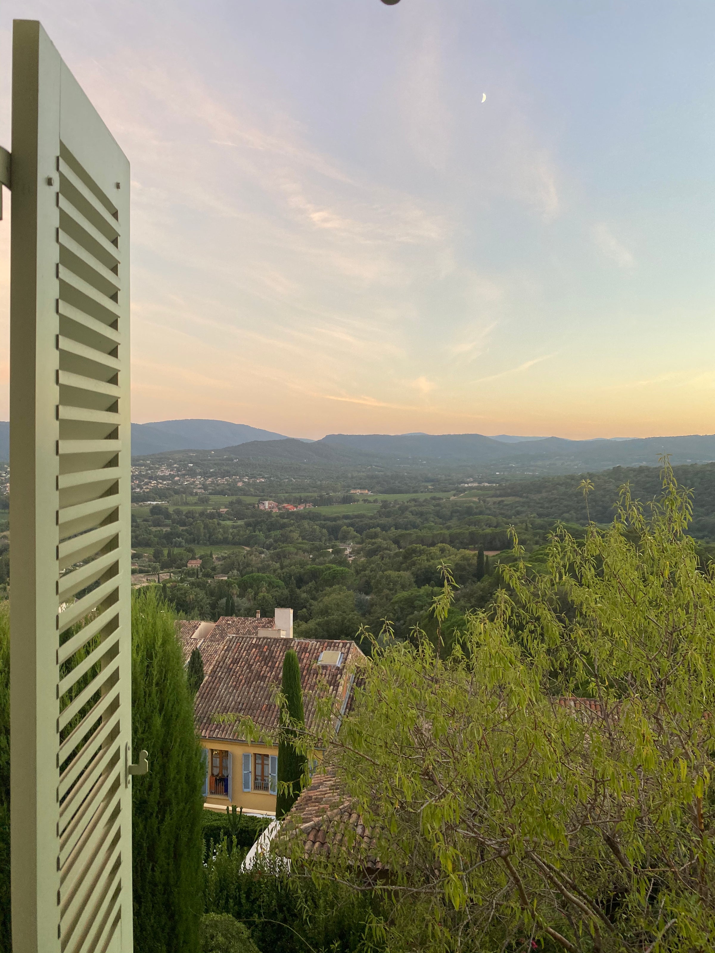 View from an open green window shutter overlooking rooftops, lush green trees, and a distant mountain range under a soft sunset sky with wispy clouds and a visible crescent moon.