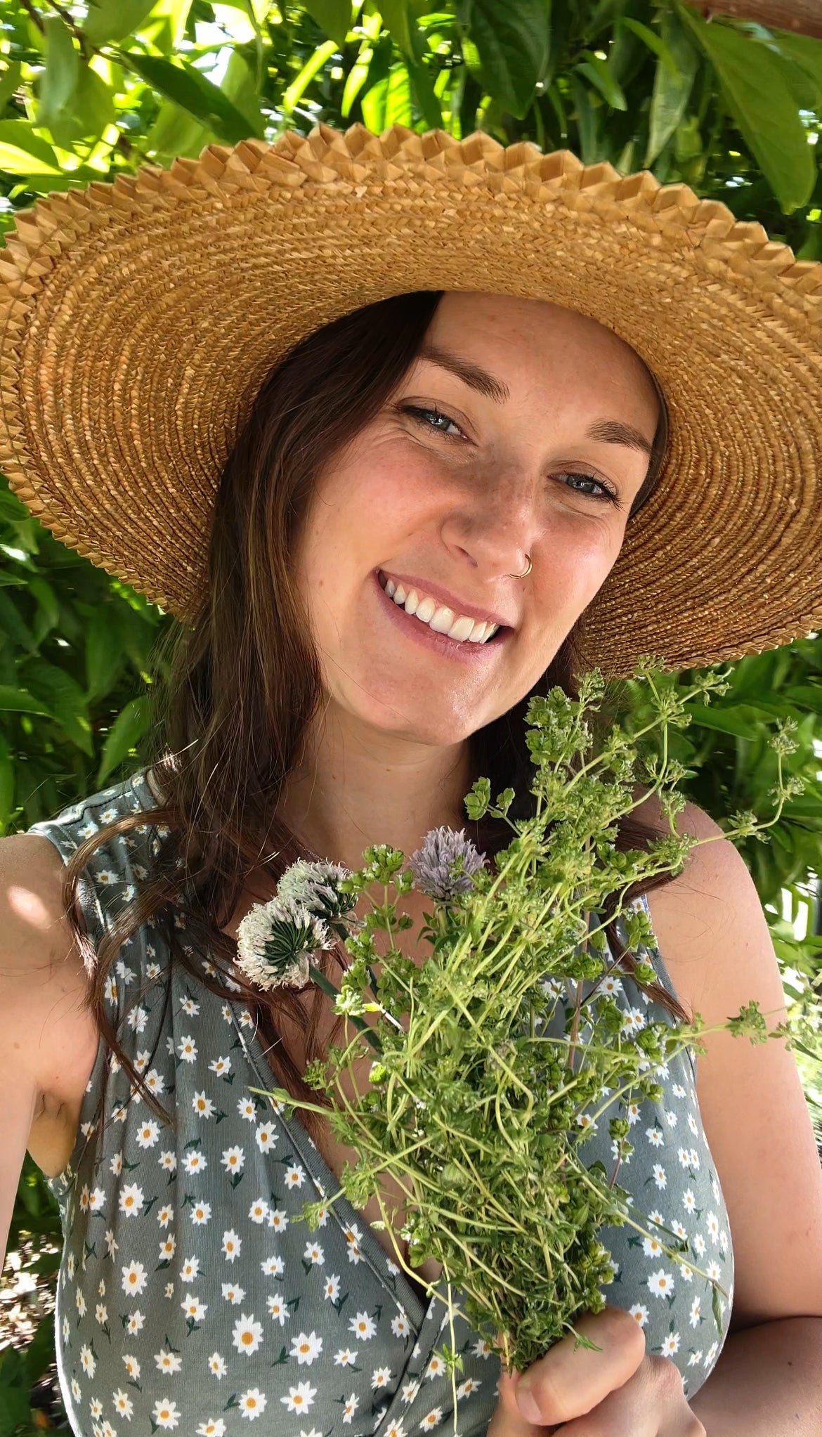 A woman wearing a wide-brimmed straw hat and a floral dress smiles while holding a bouquet of wildflowers. Green foliage and sunlight are visible in the background.