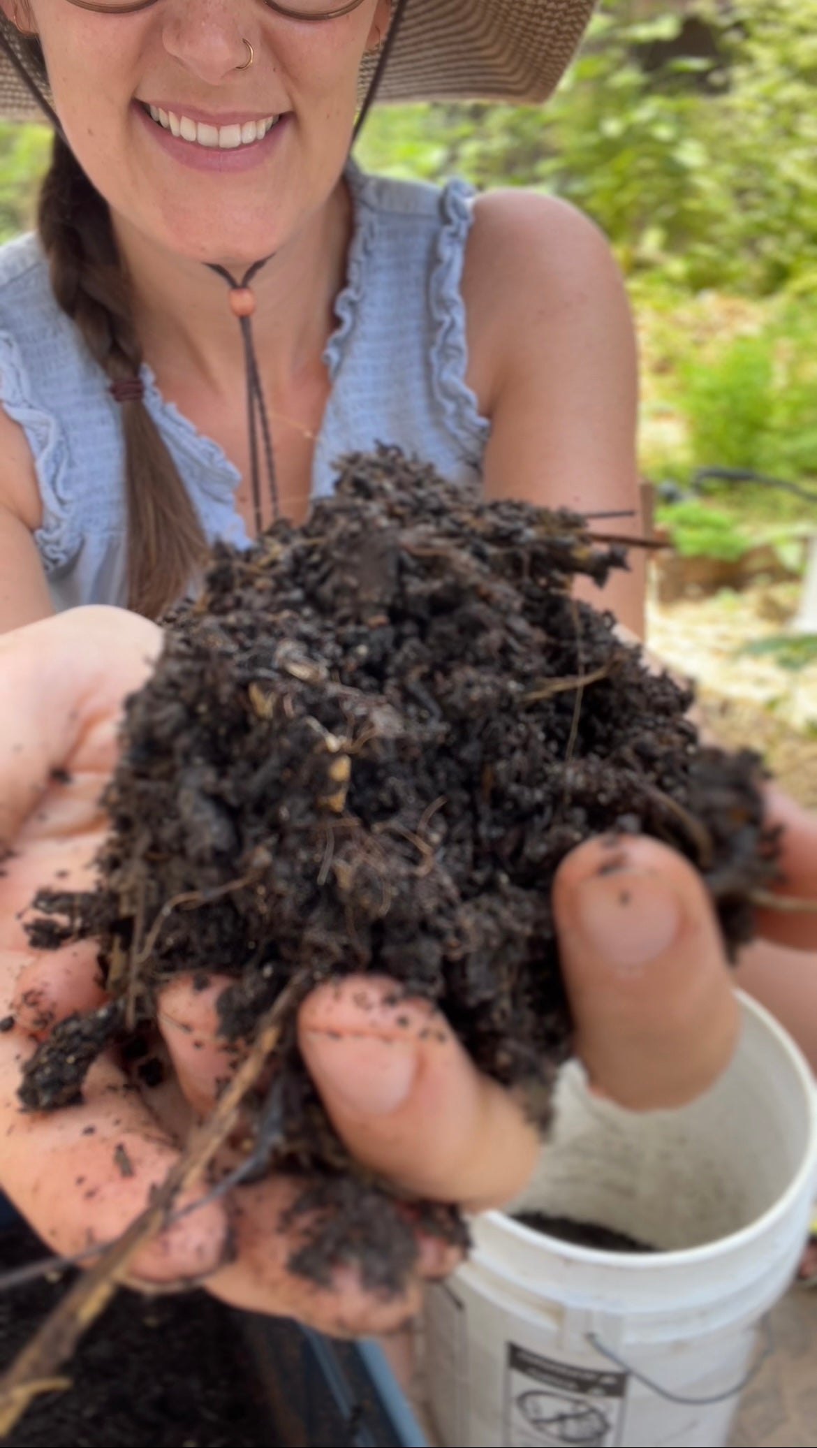 A woman outdoors holds a large handful of rich, dark soil towards the camera, smiling. She wears a sun hat, glasses, and a light blue sleeveless top, with greenery blurred in the background.