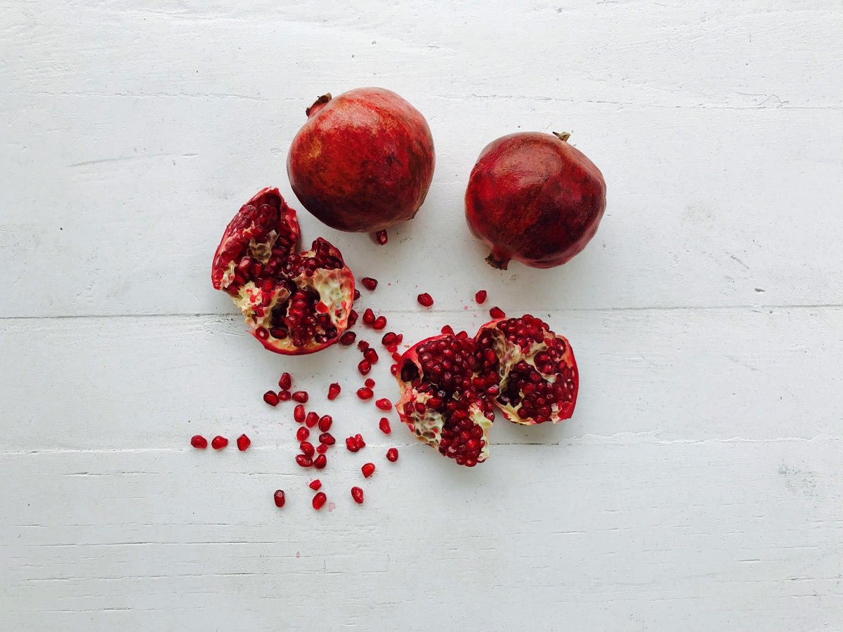 Two whole pomegranates and one pomegranate split open, with seeds scattered on a white wooden surface.