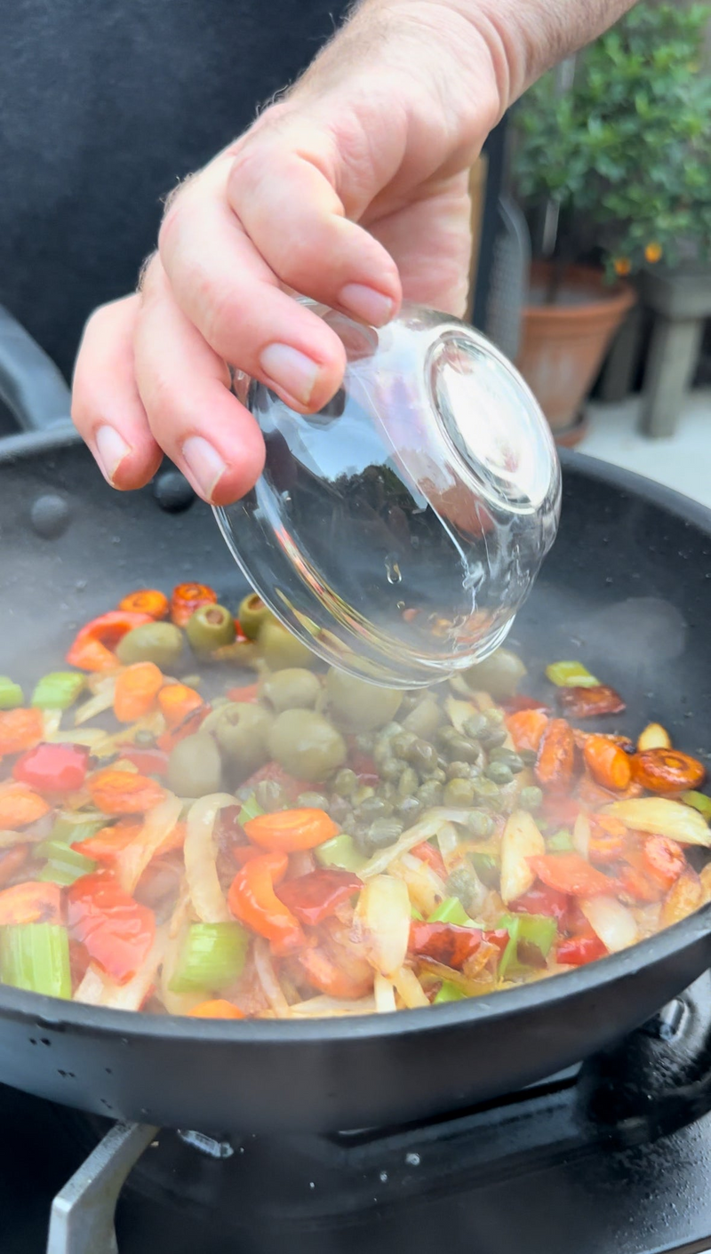 A hand pours capers from a small glass bowl into a steaming pan filled with sautéed vegetables, including tomatoes, celery, olives, and onions, outdoors.