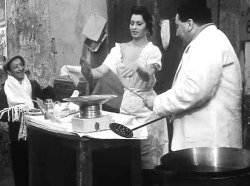 A black-and-white photo shows a woman and a man preparing food at an outdoor stall, with the woman holding dough and the man using a slotted utensil. An older person sits nearby, watching them and smiling.