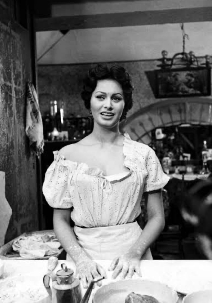 A woman in a vintage kitchen stands at a table, wearing a light-colored off-the-shoulder blouse and apron, with flour on her hands, preparing dough and smiling slightly. The background has rustic decor and kitchen items.