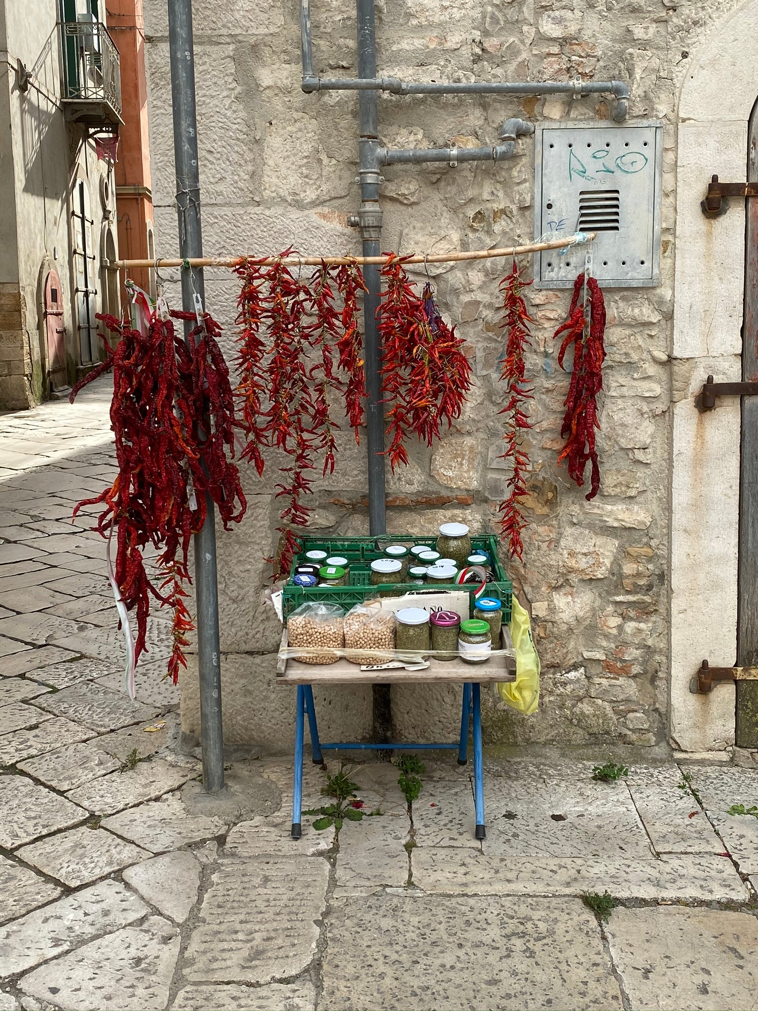 A small street market stall displays jars and containers below strings of dried red peppers hanging on a metal rod against a stone wall in an old town setting.