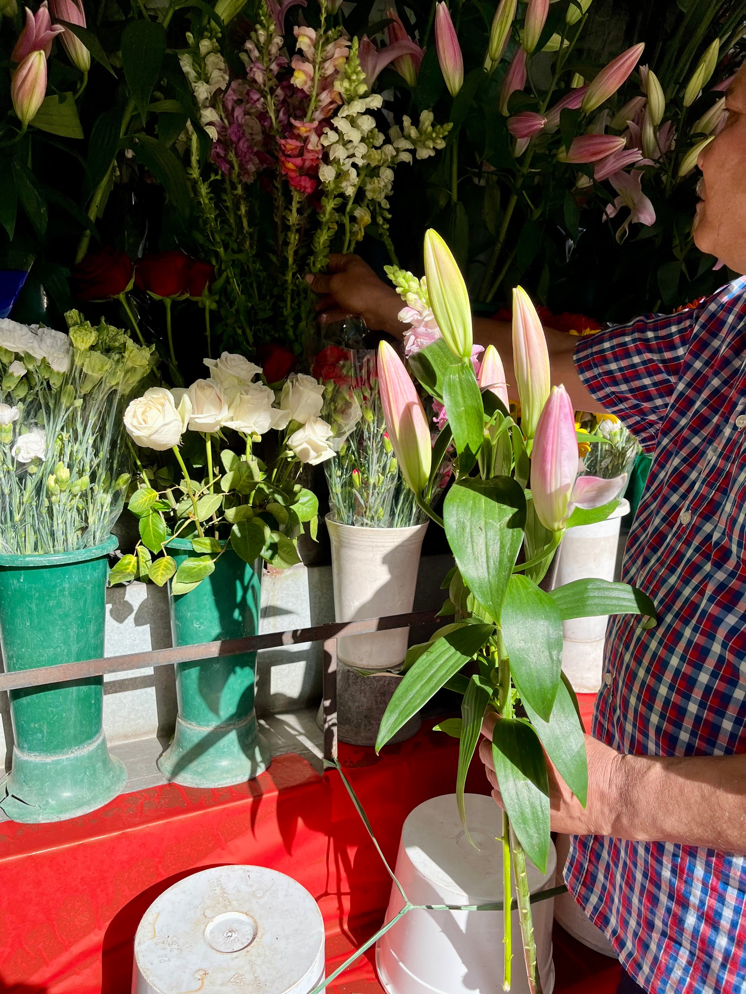 A person in a plaid shirt arranges bouquets of flowers at a market stall, holding a bunch of pink lilies. White roses and other colorful flowers are displayed in green and white vases on a red table.