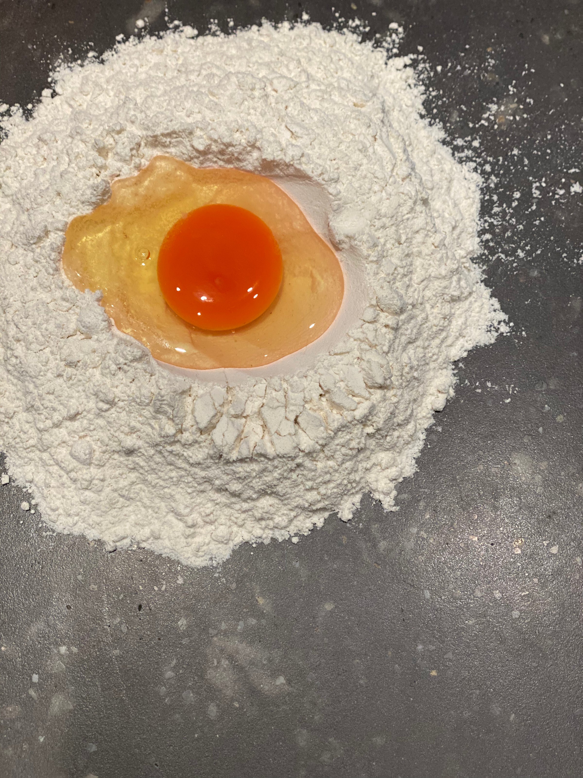 An egg cracked into a mound of flour on a dark countertop, with the flour forming a well around the egg yolk and white.