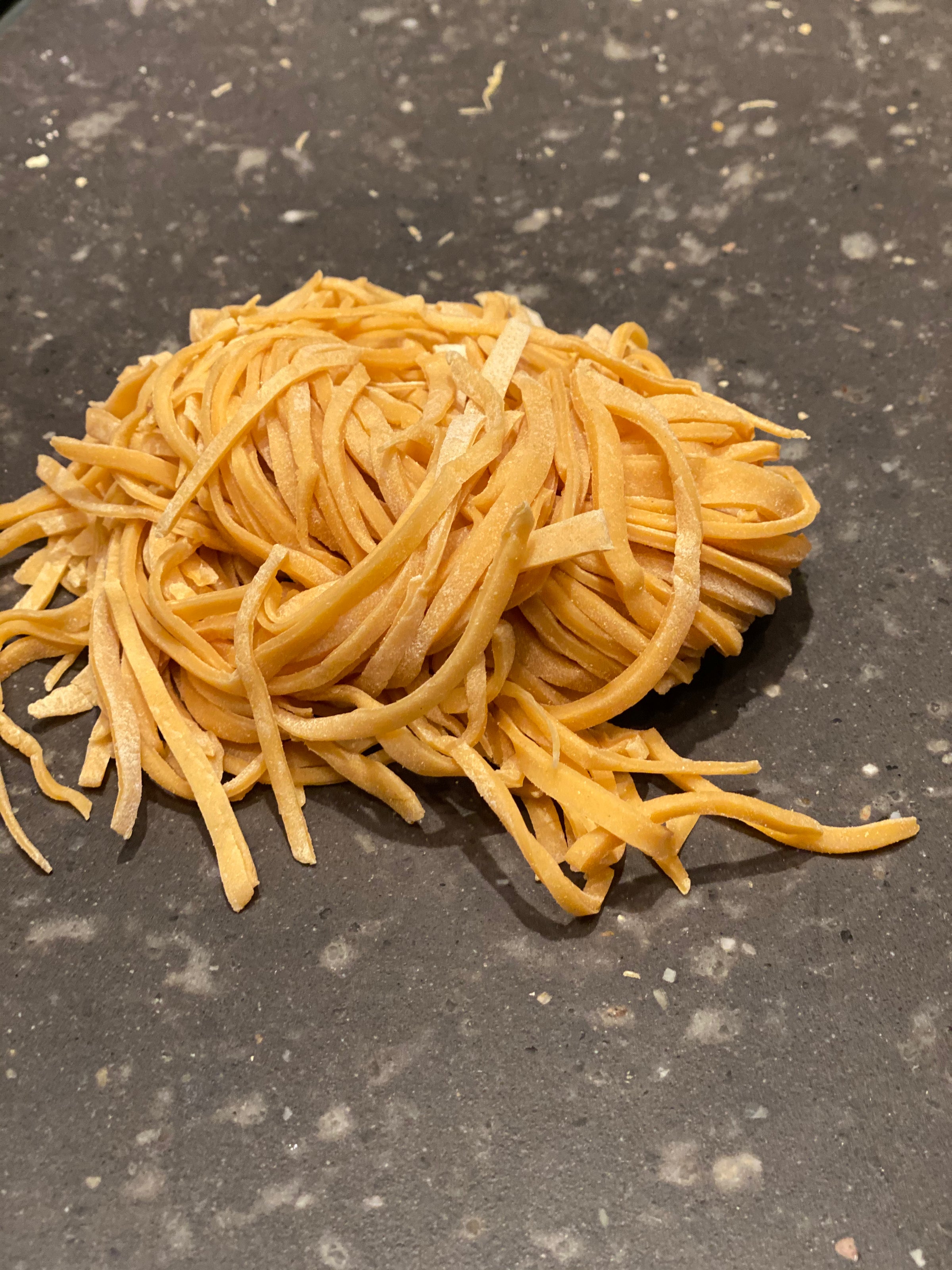 A small pile of uncooked fettuccine pasta rests on a dark, speckled countertop.