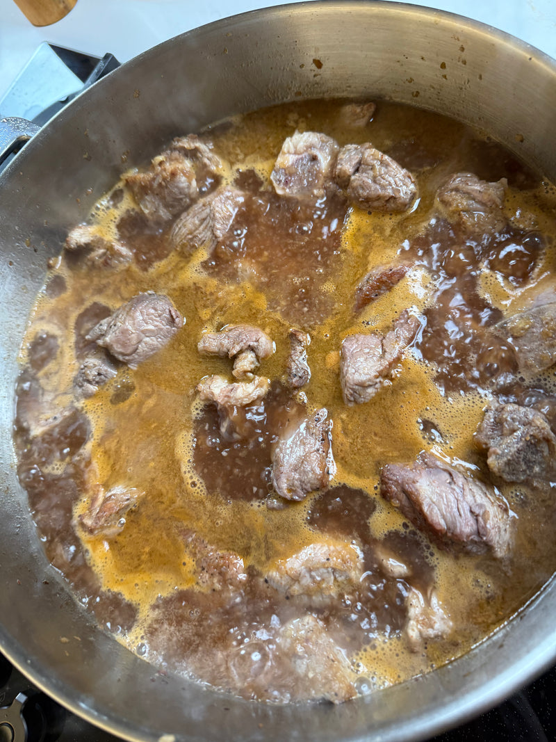 Chunks of beef simmer in a brown, bubbling broth inside a metal pot on a stovetop, with steam rising from the surface.