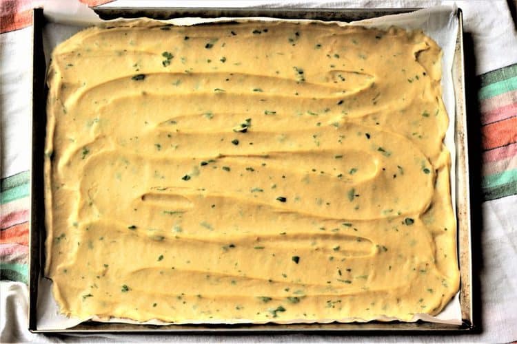 A baking tray lined with parchment paper is filled with a smooth, yellow batter mixed with green herbs, evenly spread out and ready for baking.
