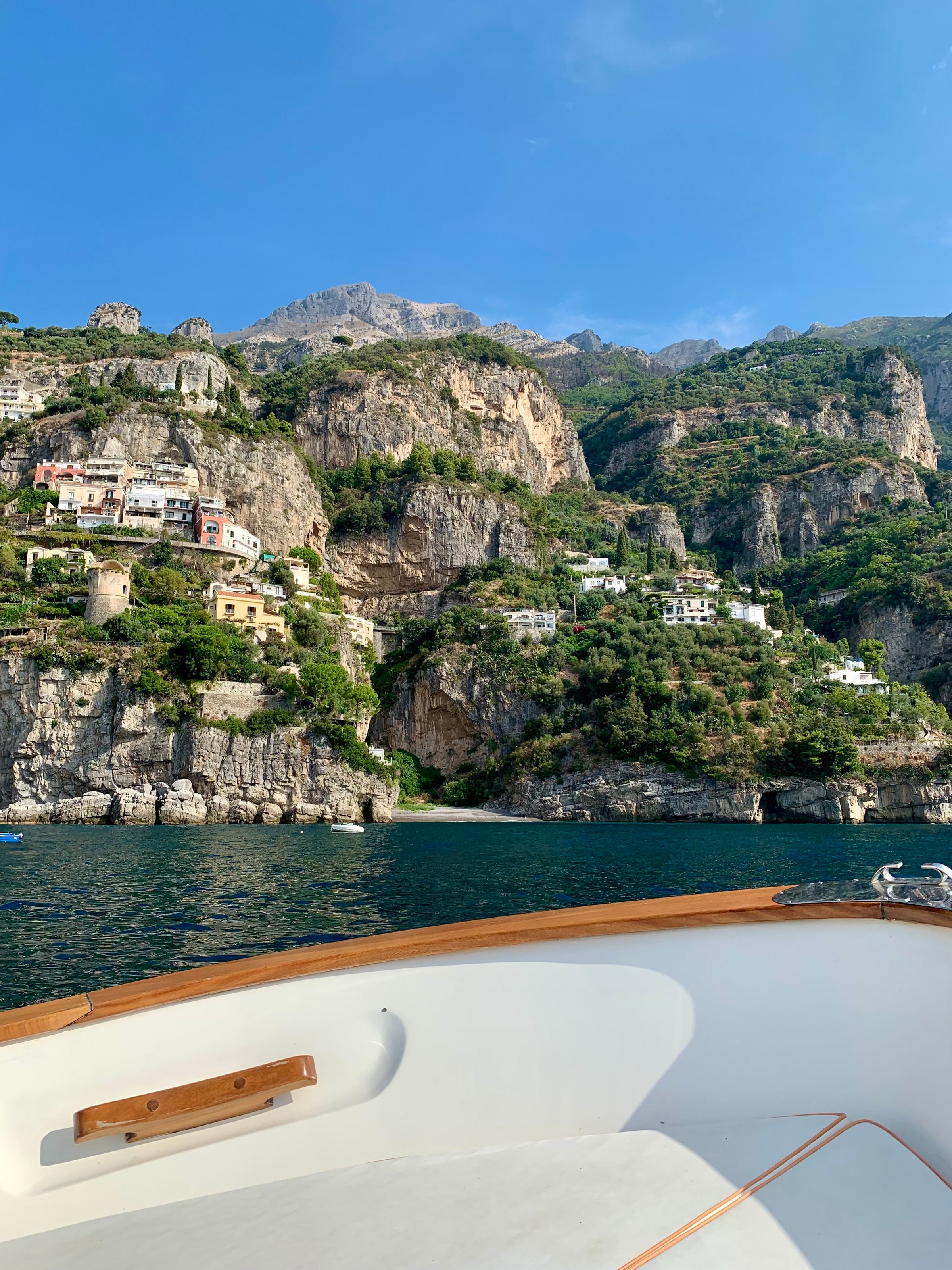 View from a boat on the water looking towards steep, rocky cliffs with scattered houses and greenery under a clear blue sky.