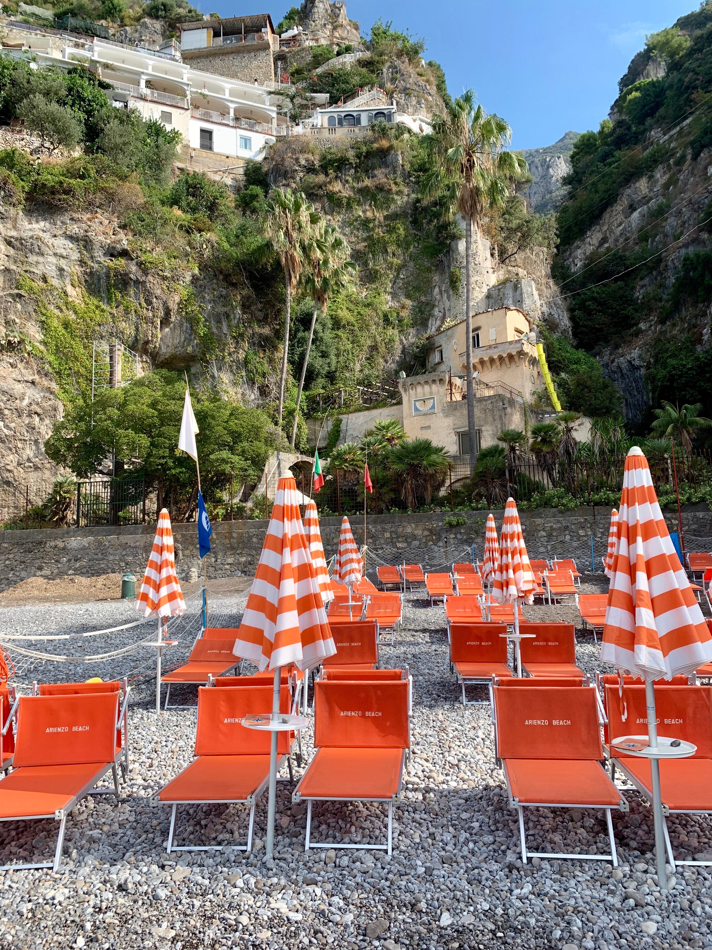 Rows of orange and white striped beach umbrellas and orange lounge chairs on a rocky beach, with steep cliffs and houses in the background under a clear blue sky.