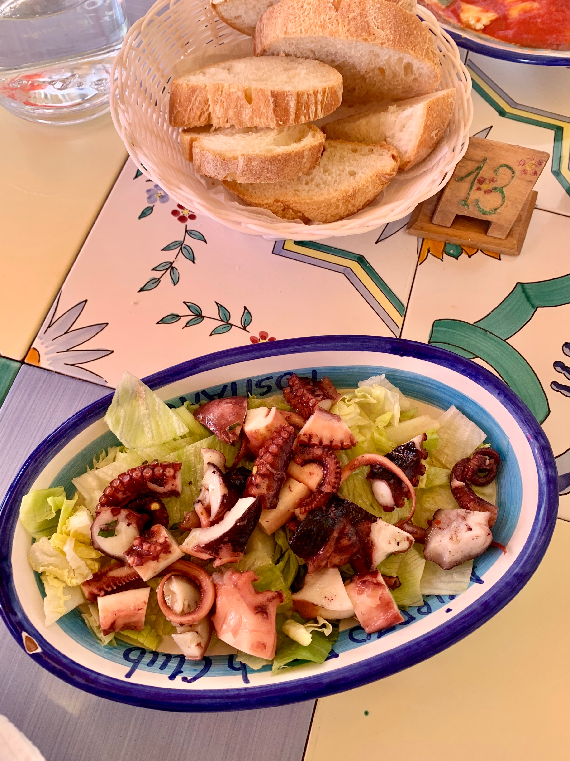 A plate of octopus salad with lettuce sits on a colorful table beside a basket of sliced bread and a glass of water. The table has a decorative floral design.