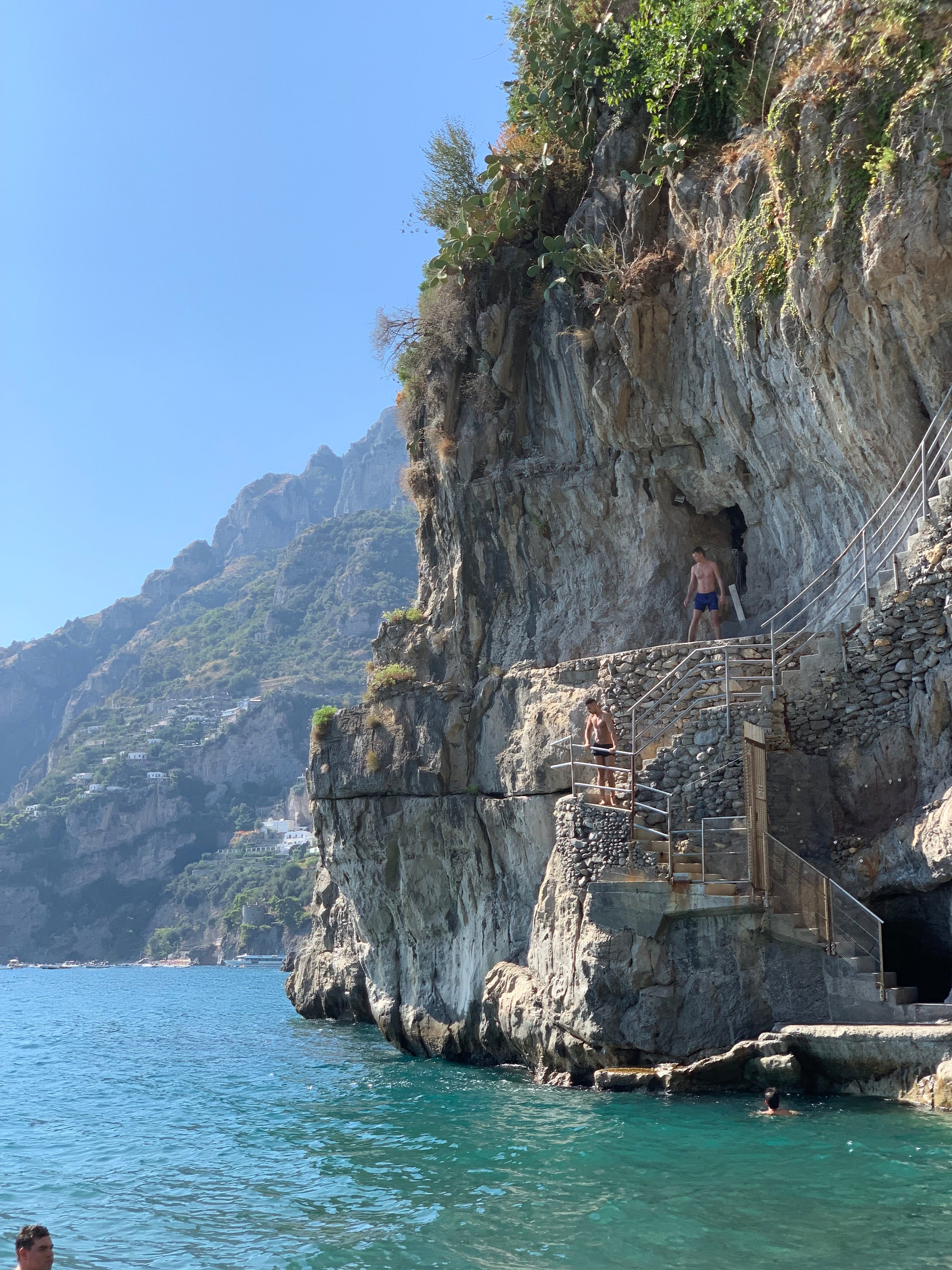 A rocky seaside cliff with stairs and railings leads down to clear blue water. People are walking on the path, and a swimmer is in the sea. Green hills and buildings are visible in the background under a clear sky.