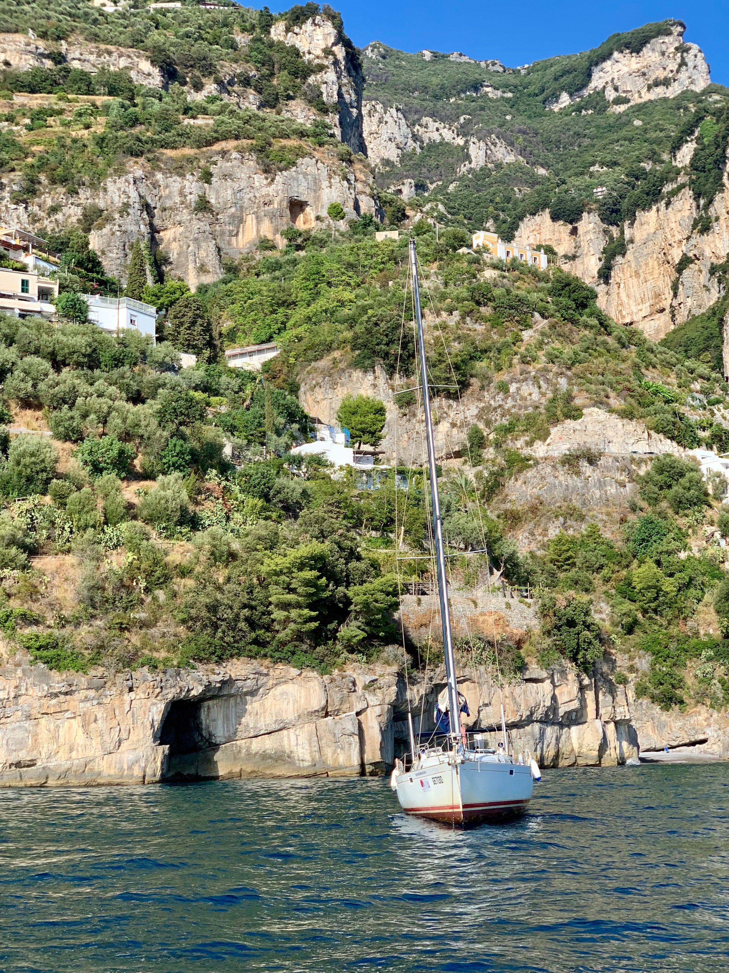 A sailboat floats on calm blue water near rocky cliffs covered with green vegetation and scattered houses, under a clear blue sky.