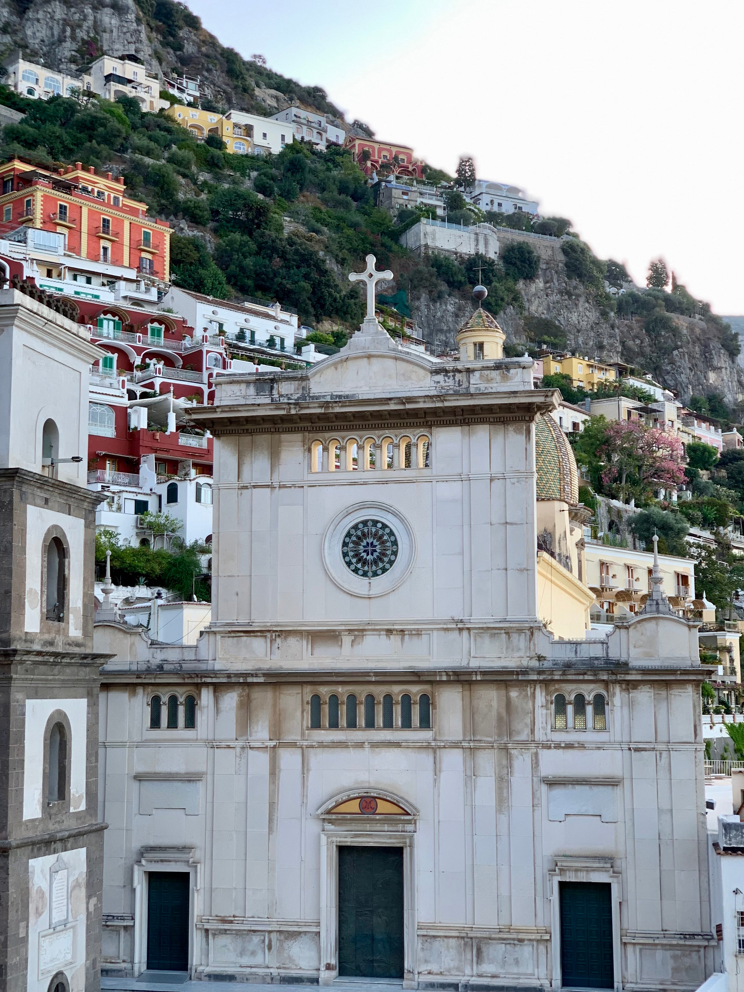 A white church with a cross on top and a circular stained glass window stands in front of colorful buildings on a steep hillside in Positano, Italy. Lush greenery and cliffs rise in the background.