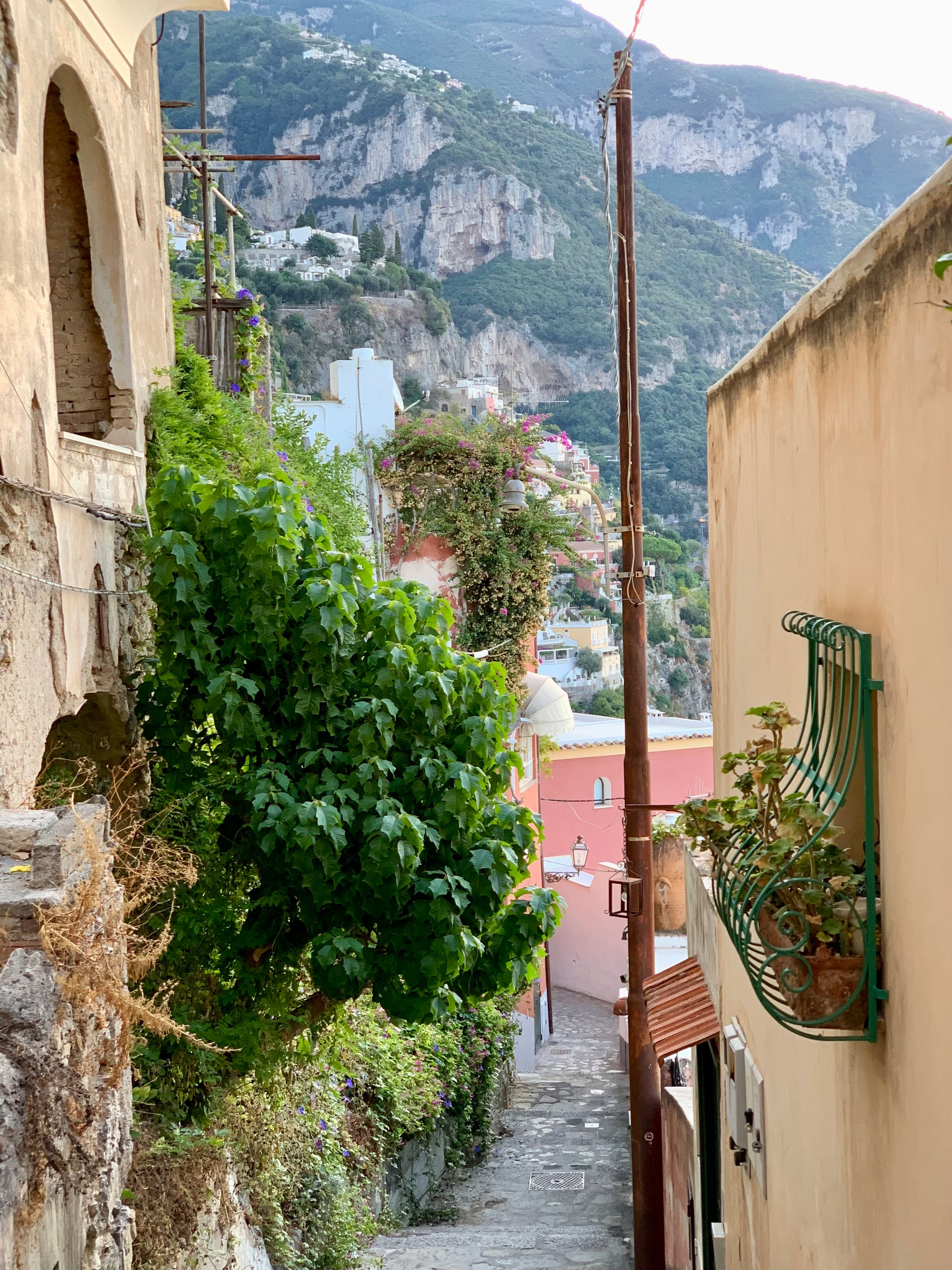 Narrow stone alleyway with colorful buildings and lush greenery, leading downhill toward distant cliffs and houses perched on steep mountainsides under a bright sky.