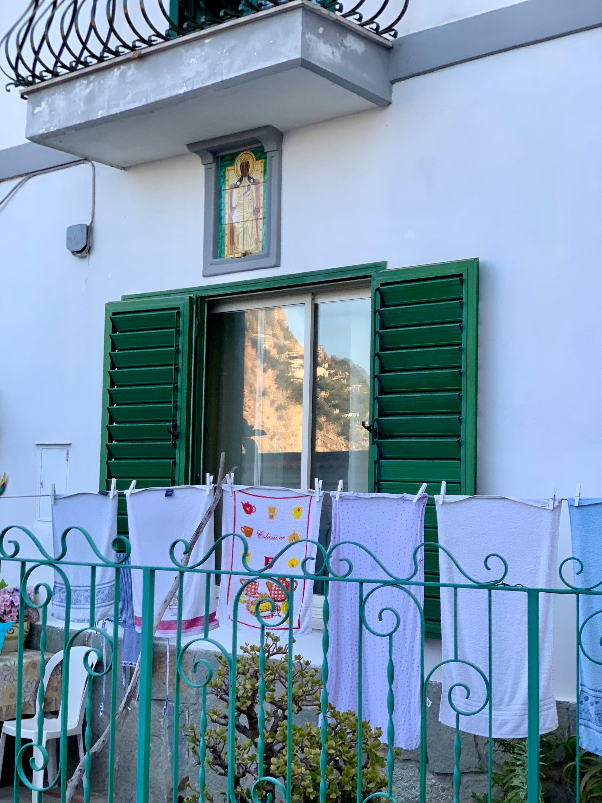 A green-shuttered window on a white building is open, with laundry hanging on a clothesline outside a green fence. There are several towels and a colorful cloth with fish and hearts on it. Shrubs are in the foreground.