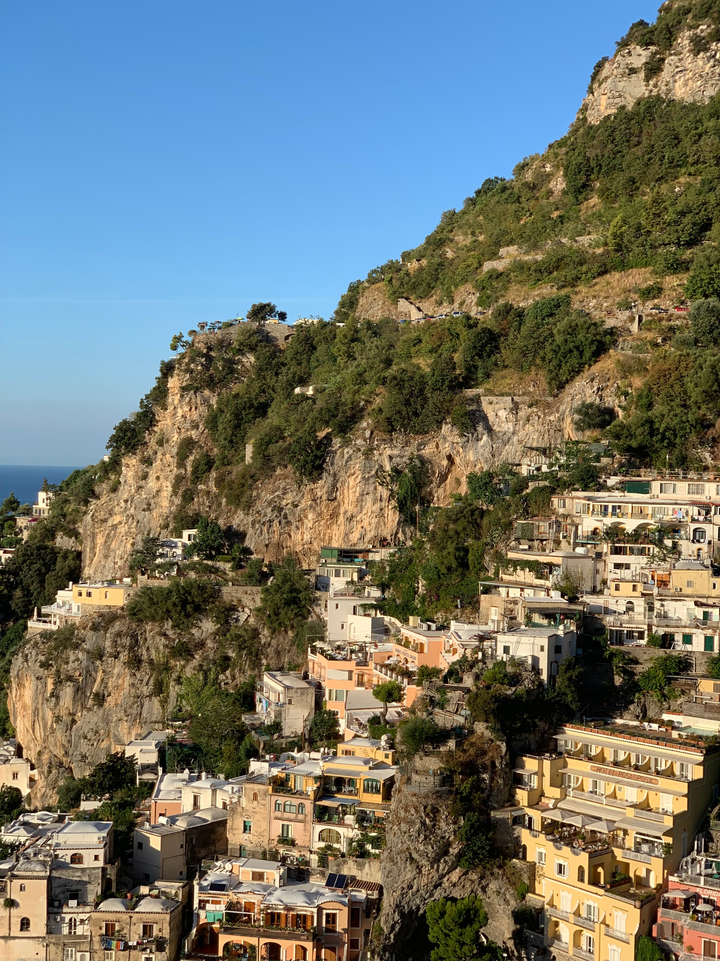 Colorful houses are built into a rocky hillside covered with lush greenery, under a clear blue sky, overlooking the sea in Positano, Italy.