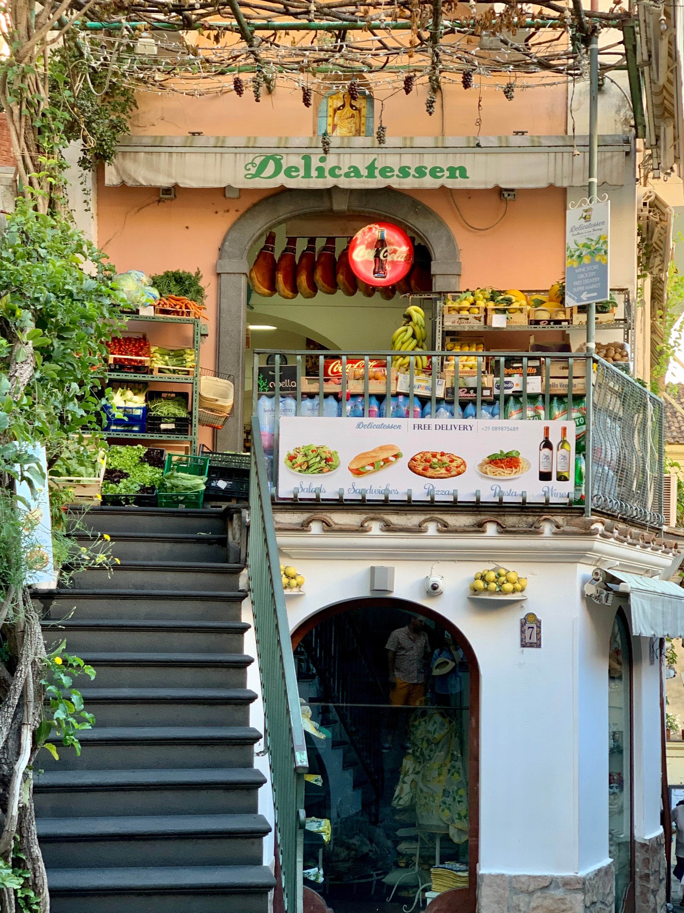 A charming delicatessen with a vine-covered staircase, fresh produce, hanging cured meats, and a balcony adorned with fruit and menu signs. The building has a rustic, inviting façade.