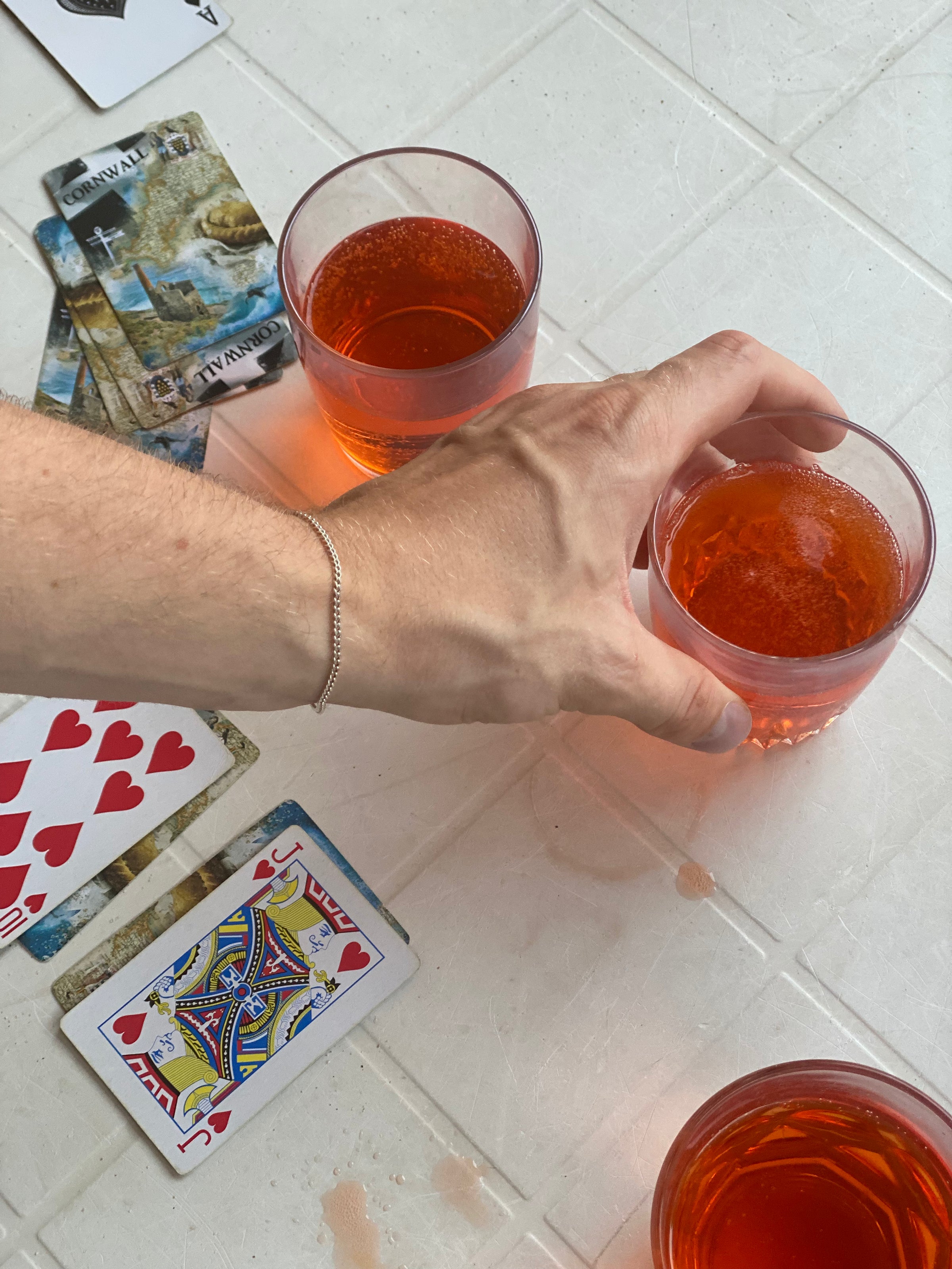 A hand reaches for a glass of orange-red drink on a white table scattered with playing cards, tarot cards, and other similar drinks.