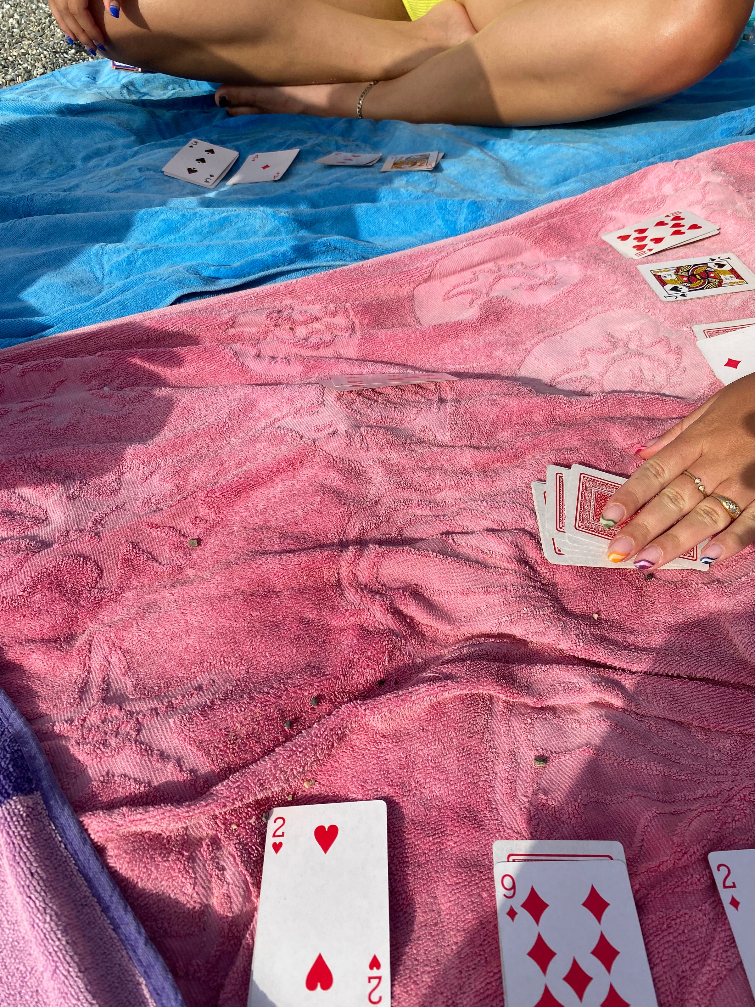 Two people playing cards on colorful towels outdoors; one persons crossed legs and the other’s hand holding playing cards are visible, with several cards laid out on the towels.