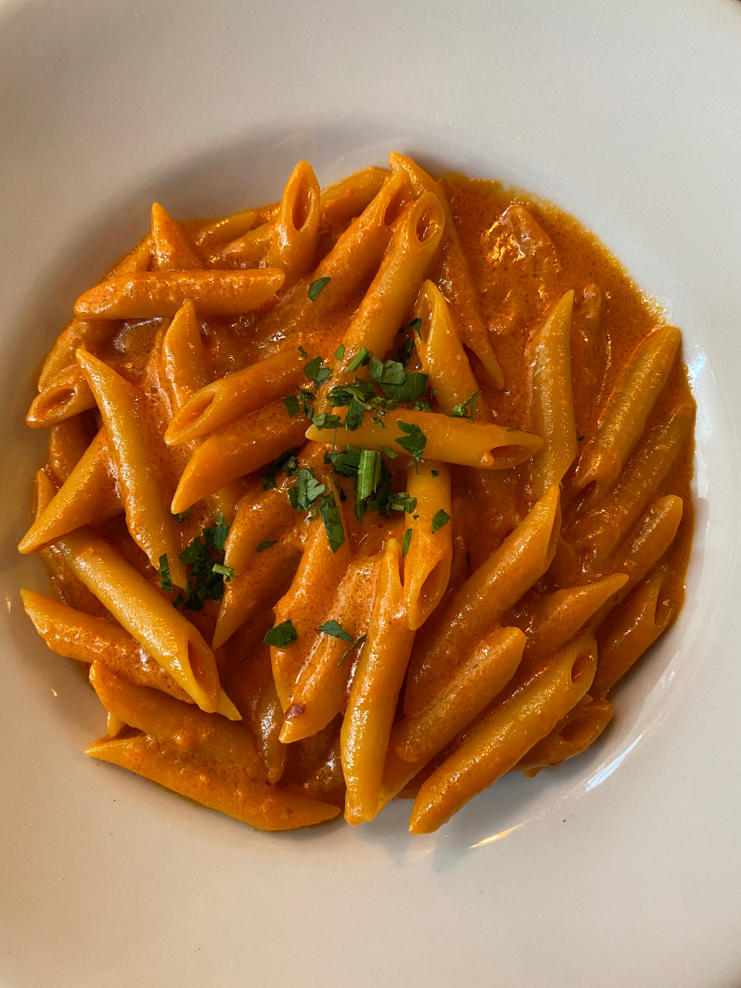 A white plate filled with penne pasta coated in a creamy tomato sauce, garnished with chopped fresh herbs.
