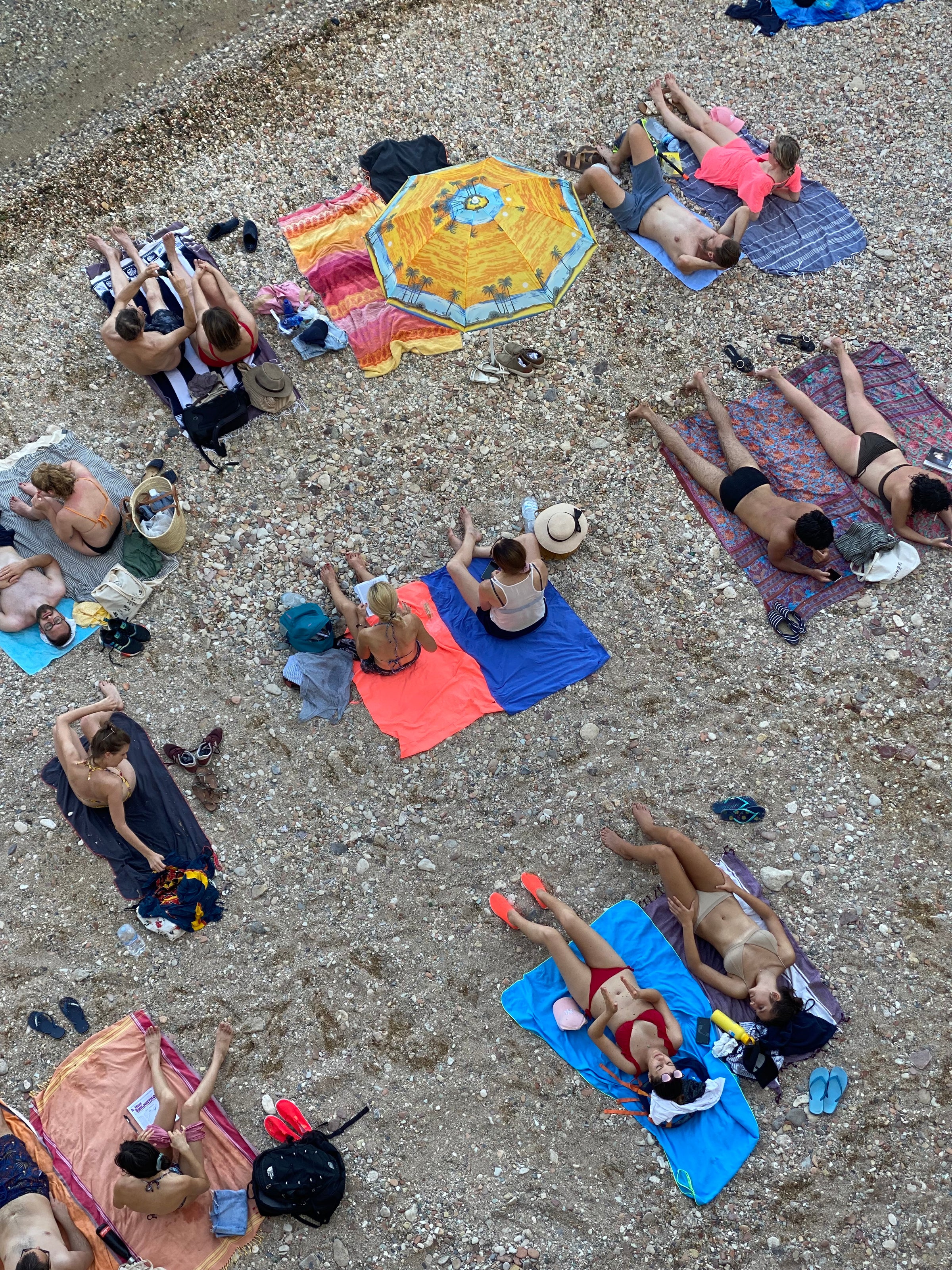 Aerial view of people relaxing on a pebble beach, lying on colorful towels and blankets, some under a yellow umbrella. Most are wearing swimsuits, and various bags and shoes are scattered around.
