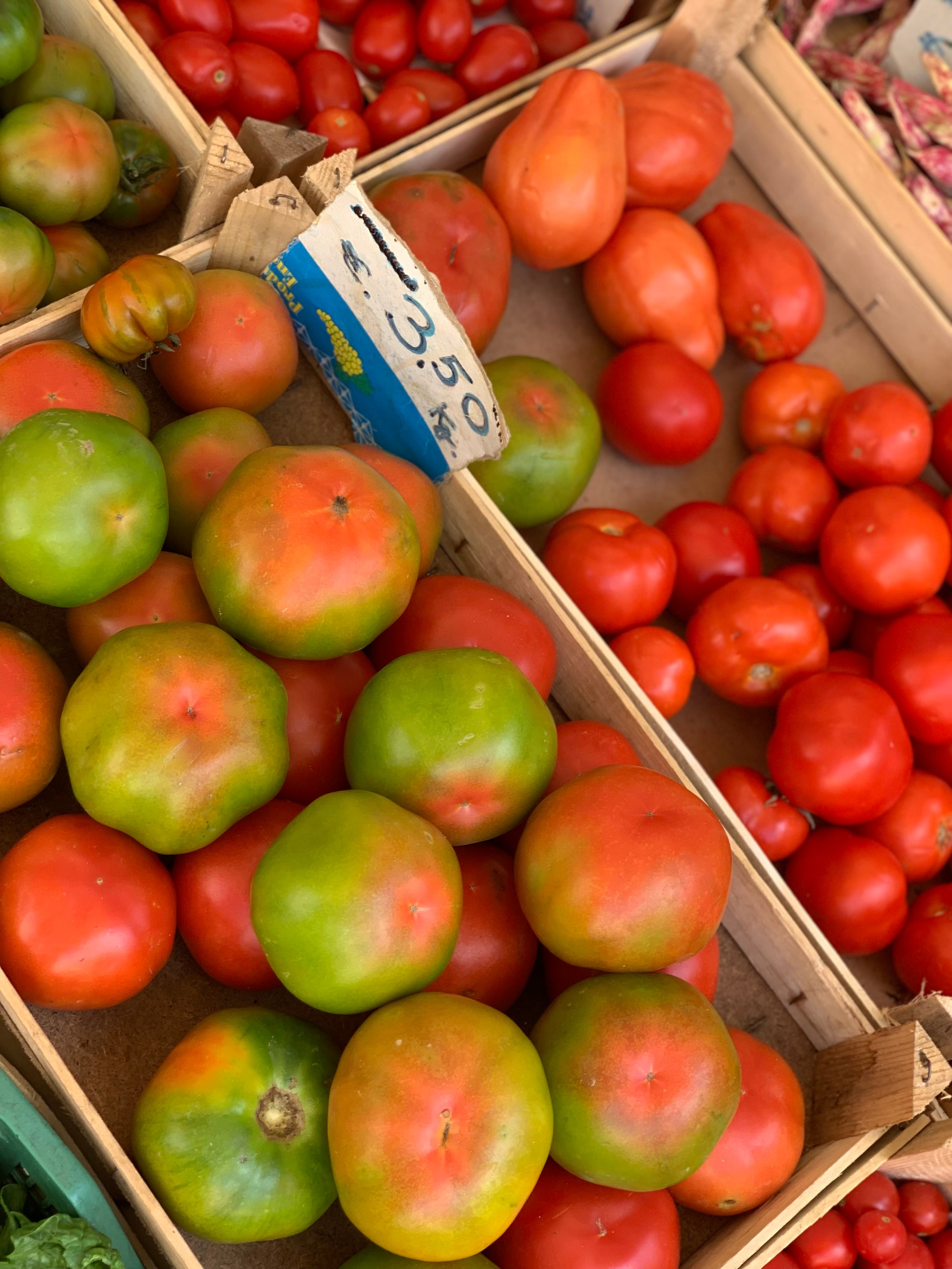 Heirloom tomatoes with red and green skin displayed in a wooden crate at a market, surrounded by other varieties of red tomatoes. A handwritten price sign reads “3.50€/kg”.