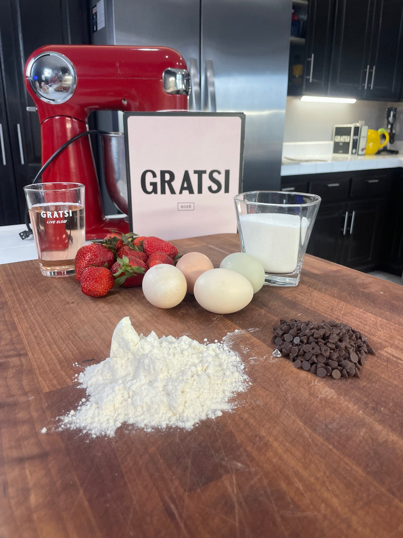 A wooden kitchen counter displays flour, chocolate chips, eggs, strawberries, sugar, two glasses, and a red stand mixer, with a GRATSI sign in the background and a modern kitchen setting.