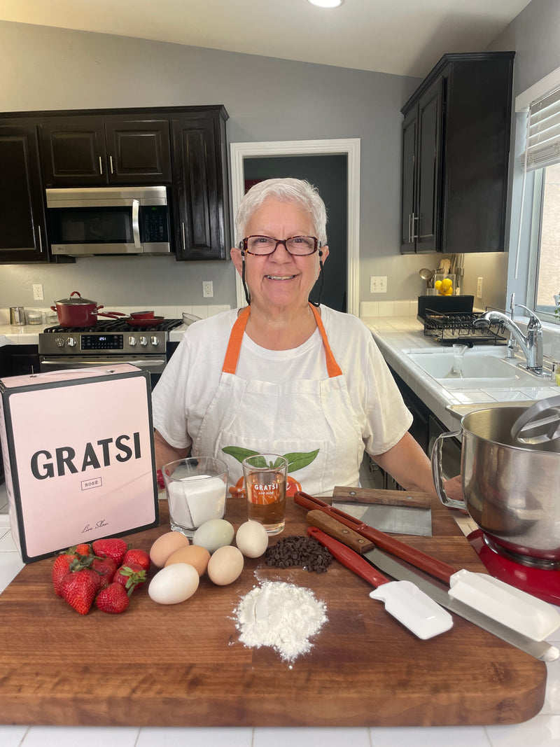 An older woman with short white hair and glasses smiles in a kitchen, wearing an apron. In front of her are baking ingredients: eggs, strawberries, flour, chocolate chips, sugar, utensils, a Gratsi box, and a stand mixer.