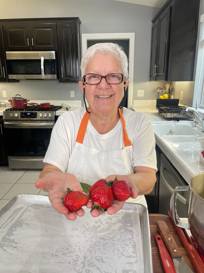 An older woman wearing glasses and an apron stands in a kitchen, smiling and holding several fresh strawberries in her outstretched hands over a baking sheet.