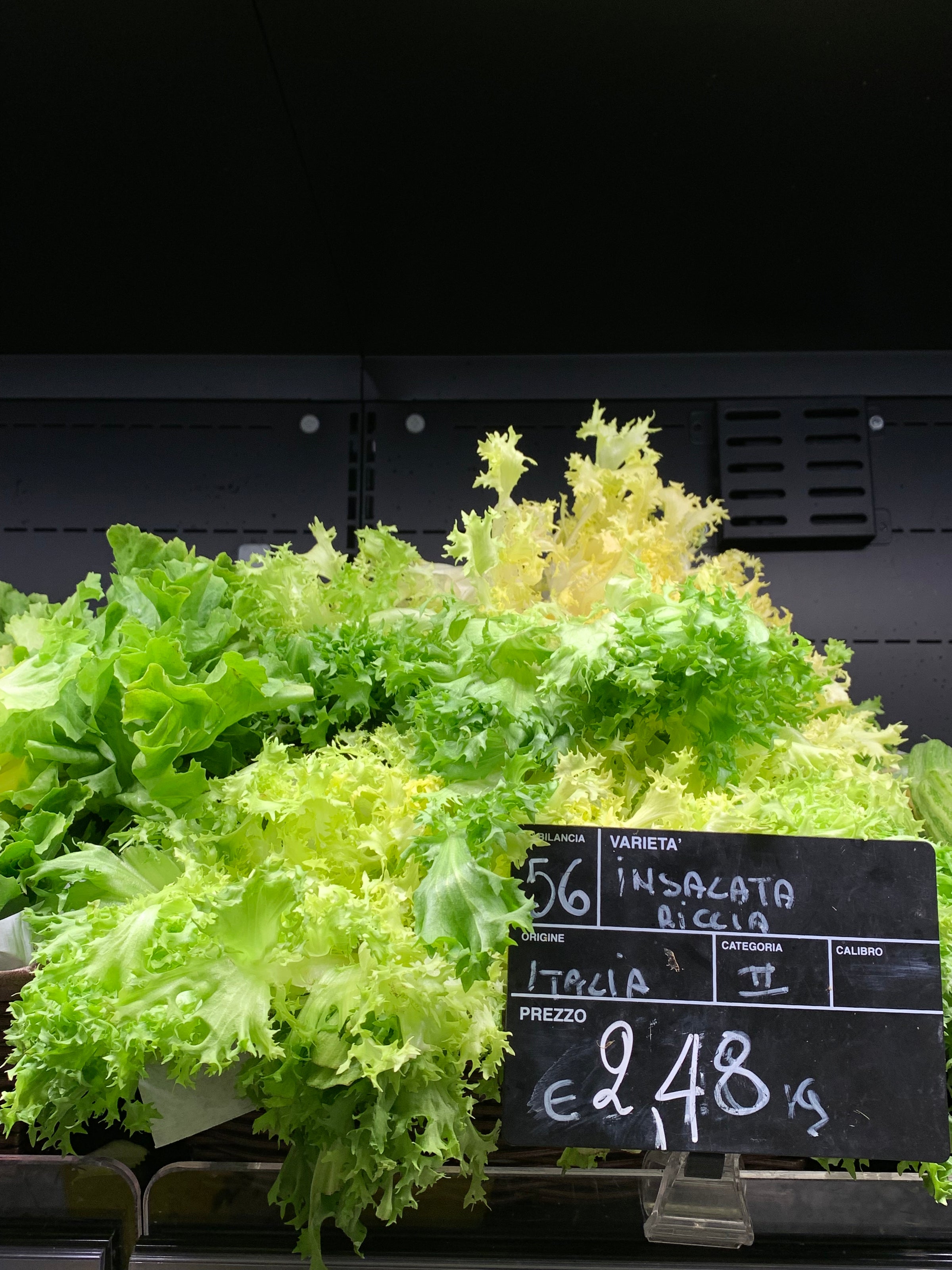 A pile of fresh green curly lettuce displayed in a supermarket. A black price sign in Italian indicates Insalata Riccia from Italy, priced at €2.48 per kilogram.