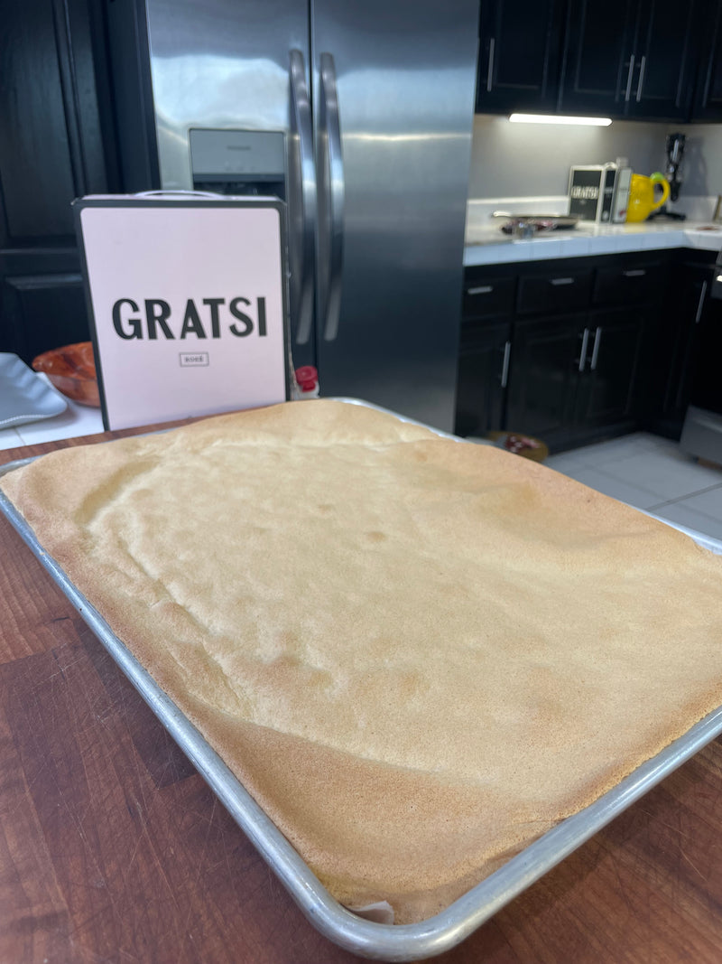 A large baking sheet filled with smooth, freshly baked cake sits on a wooden countertop in a kitchen. In the background, there’s a box labeled “GRATSI” near a stainless steel refrigerator and black cabinets.