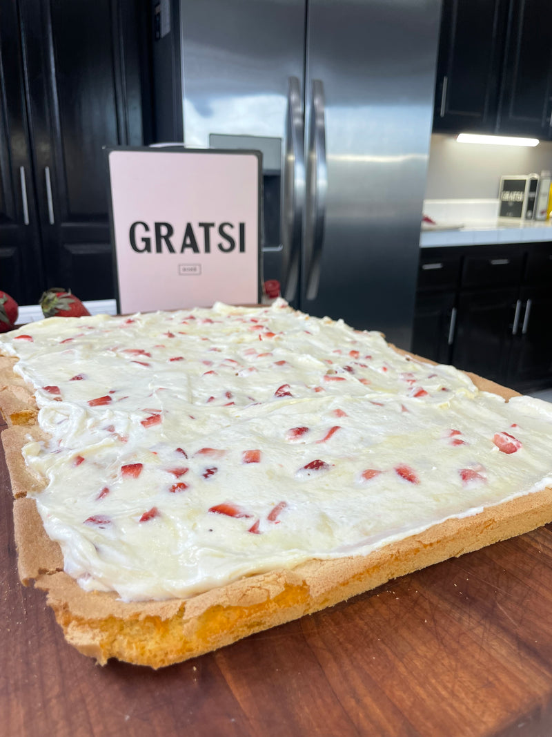A sheet cake topped with white frosting and pieces of strawberries sits on a wooden surface. In the background, there is a sign that says GRATSI and a modern kitchen with dark cabinets and a stainless steel fridge.