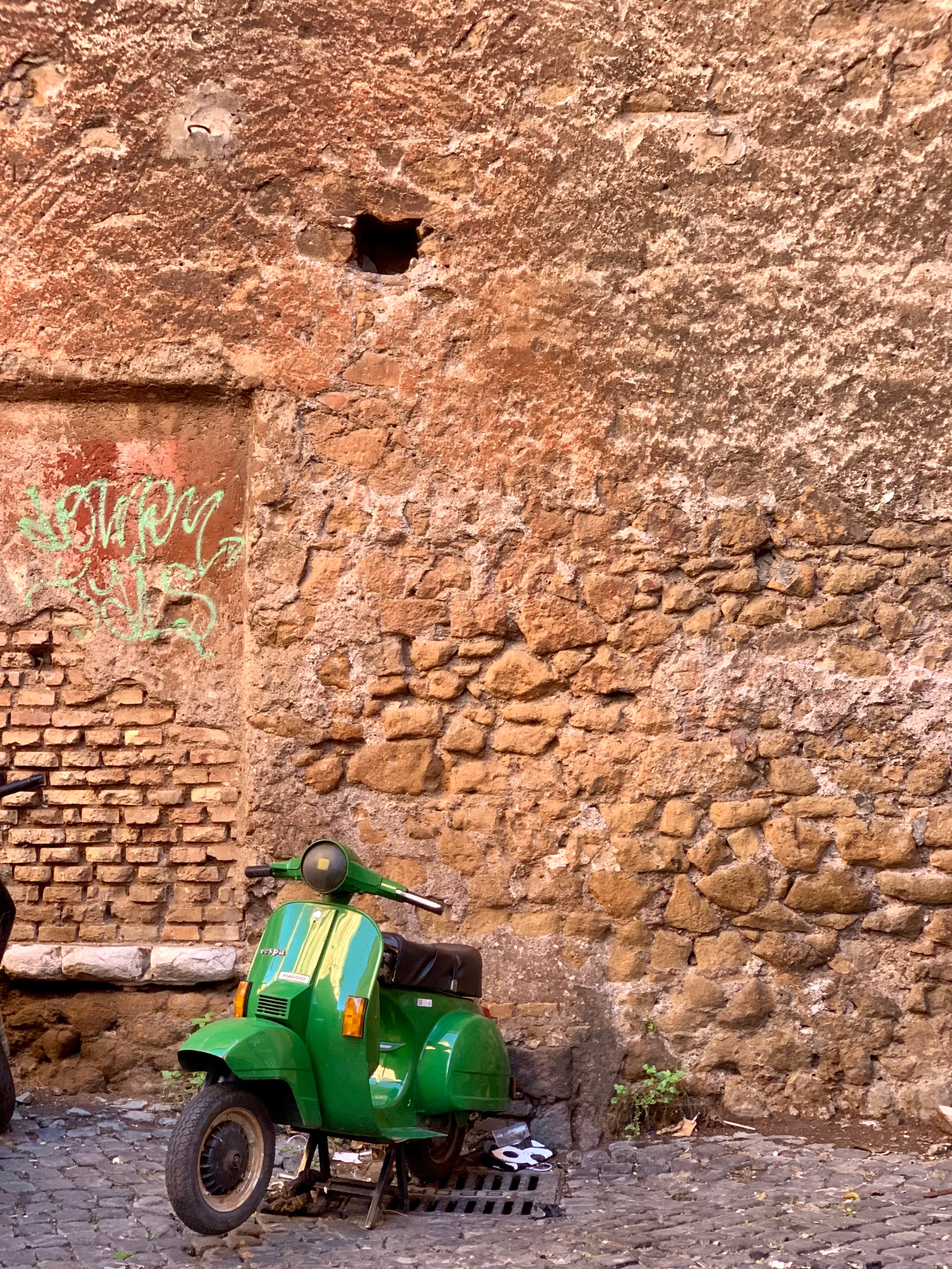 A bright green scooter is parked against an old, textured stone wall with faded graffiti and a small hole near the top, on a cobblestone street.