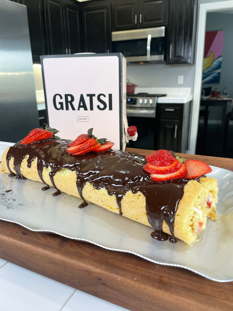 A rolled sponge cake topped with chocolate glaze and sliced strawberries sits on a platter. Behind it is a Gratsi-branded box in a modern kitchen with dark cabinets and a stainless steel oven.