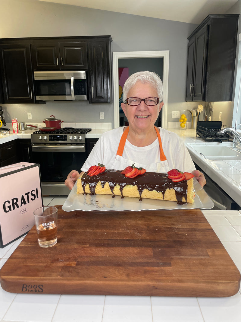 A smiling older woman with short white hair and glasses holds a large cake topped with chocolate glaze and strawberries in a bright kitchen. A pink GRATS! box and a filled shot glass are on a wooden counter in front of her.