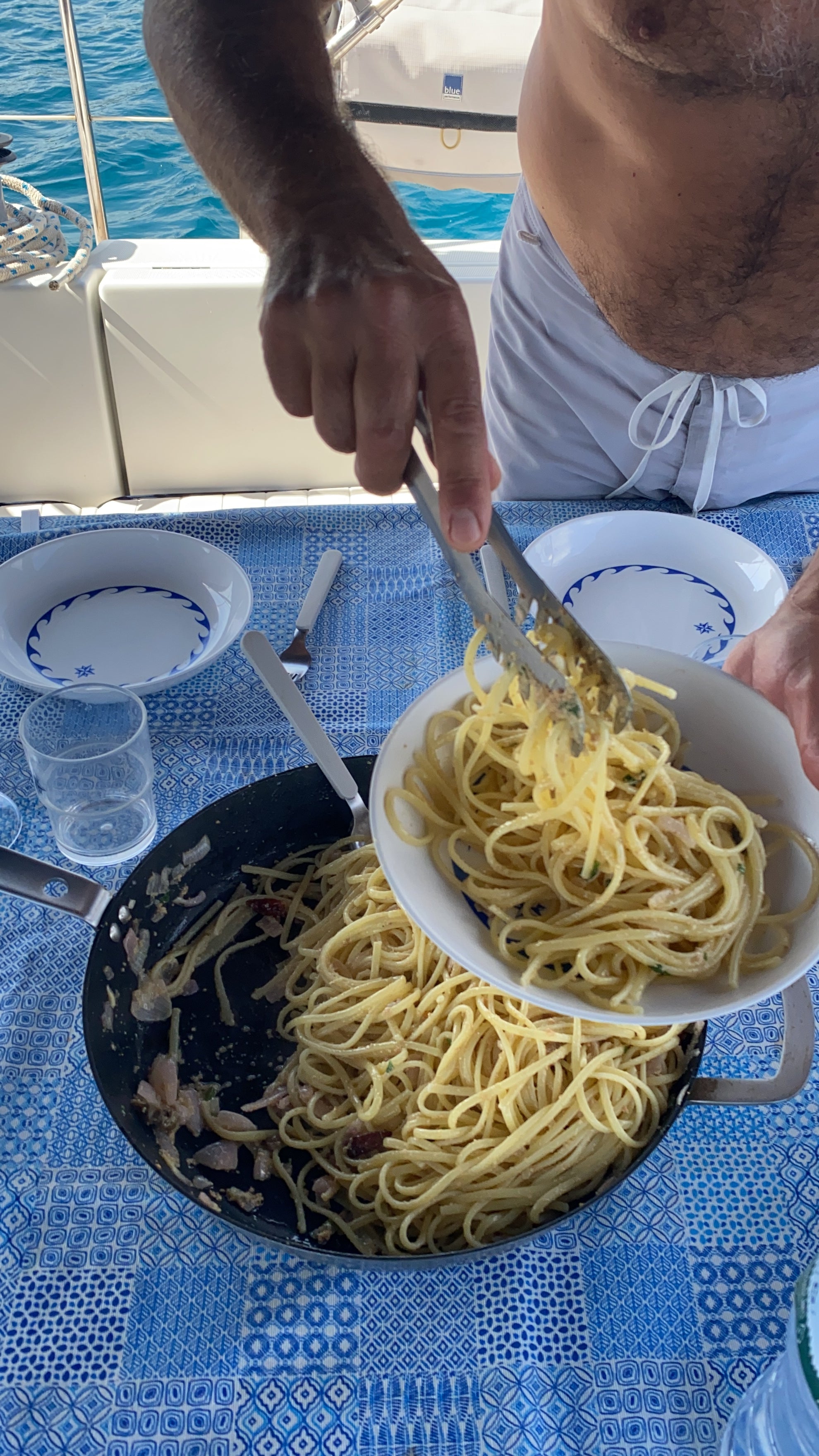 A person in swimwear serves spaghetti from a pan into a bowl on a blue-patterned tablecloth set with plates, cutlery, and a plastic cup, with a blurred view of water and a boat in the background.