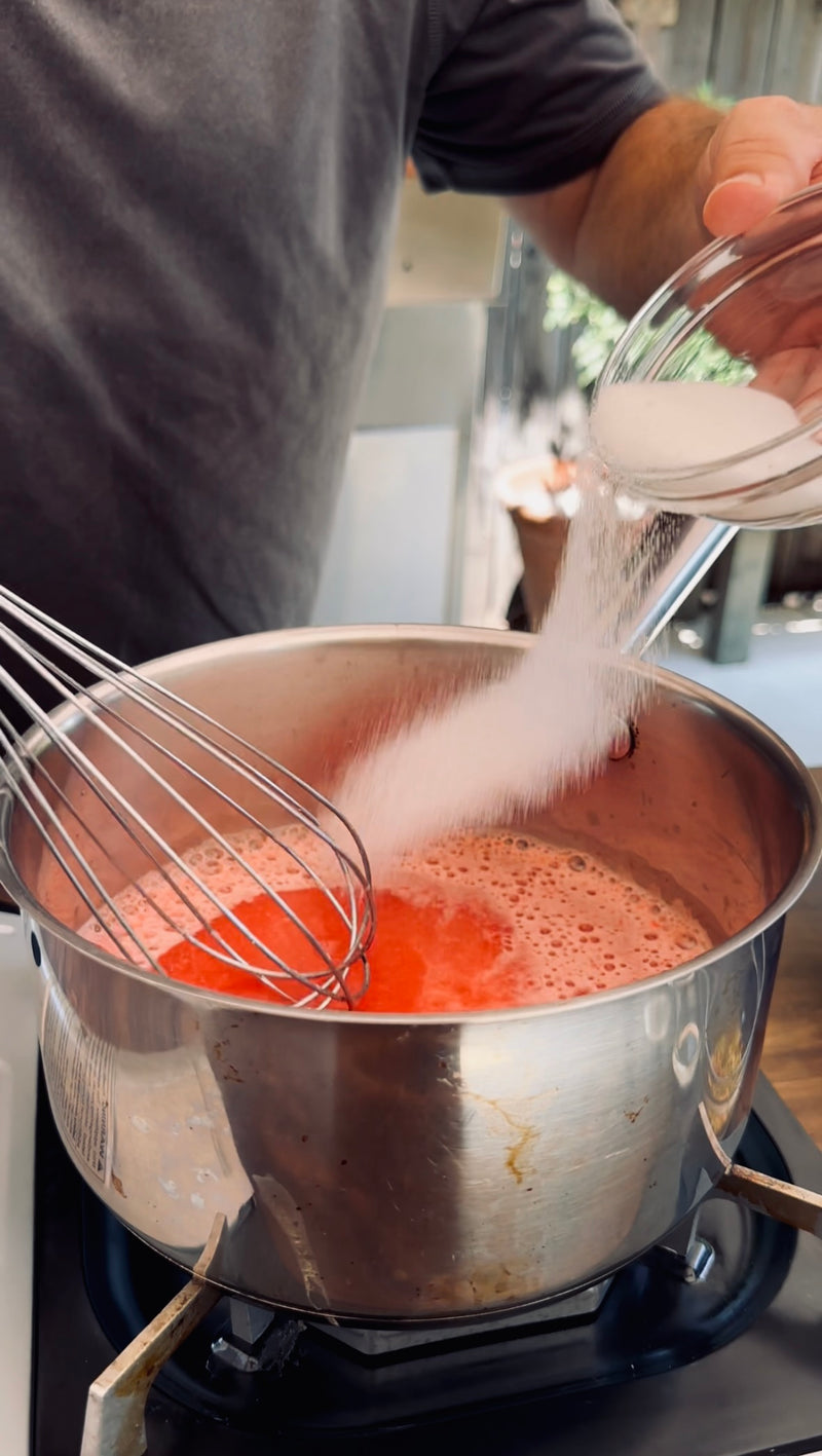 A person pours sugar from a bowl into a pot of red liquid while whisking on a stovetop.