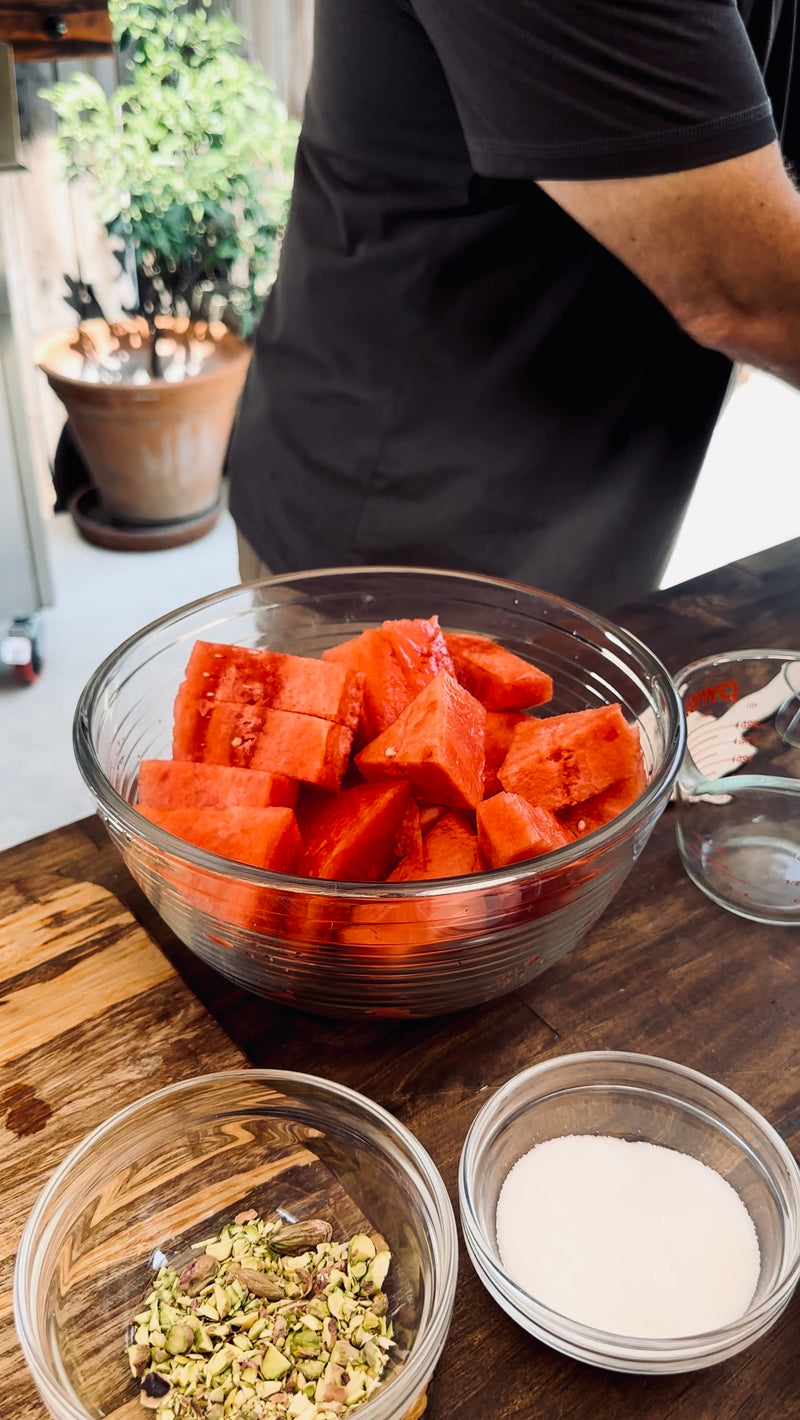A glass bowl filled with cubed watermelon sits on a wooden table, surrounded by small bowls containing sugar and chopped pistachios. A person stands nearby, partially out of frame.