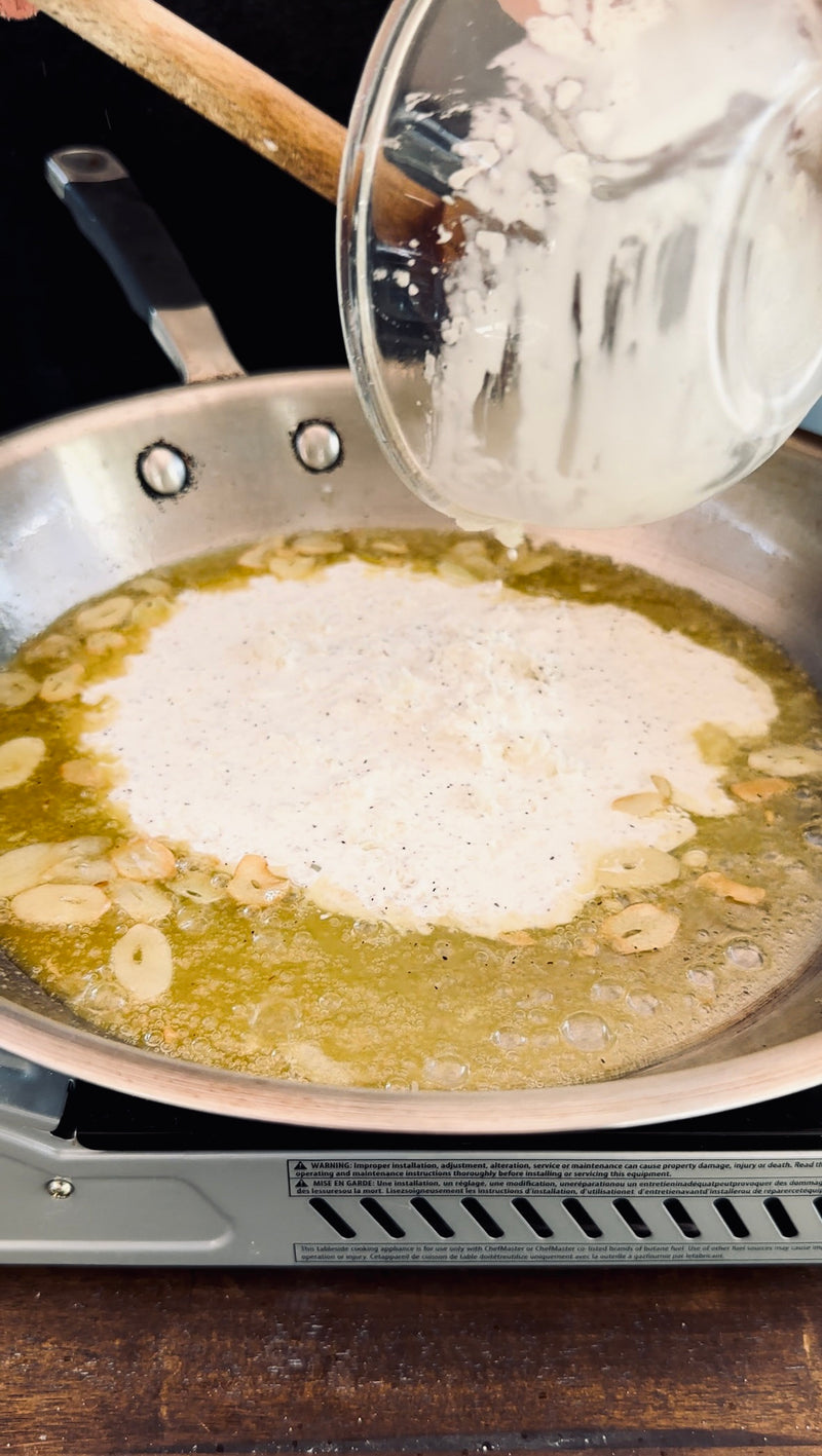 Sliced garlic cooking in melted butter in a pan, while a creamy white mixture is being poured in from a glass bowl above.