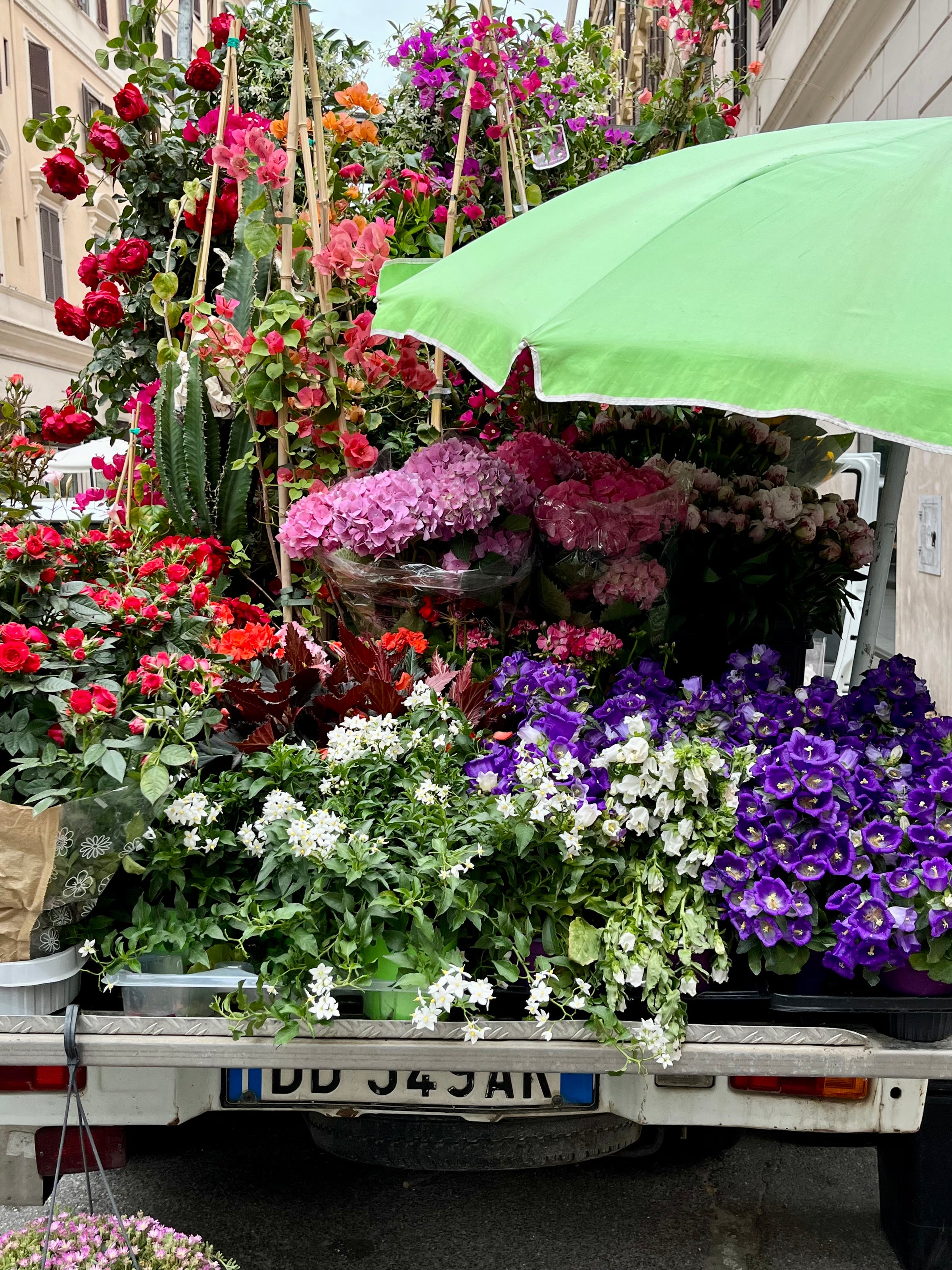 A truck loaded with colorful flowers—roses, hydrangeas, petunias, and more—parked on a city street. A large green umbrella shades some of the blooms, creating a vibrant, lush display.